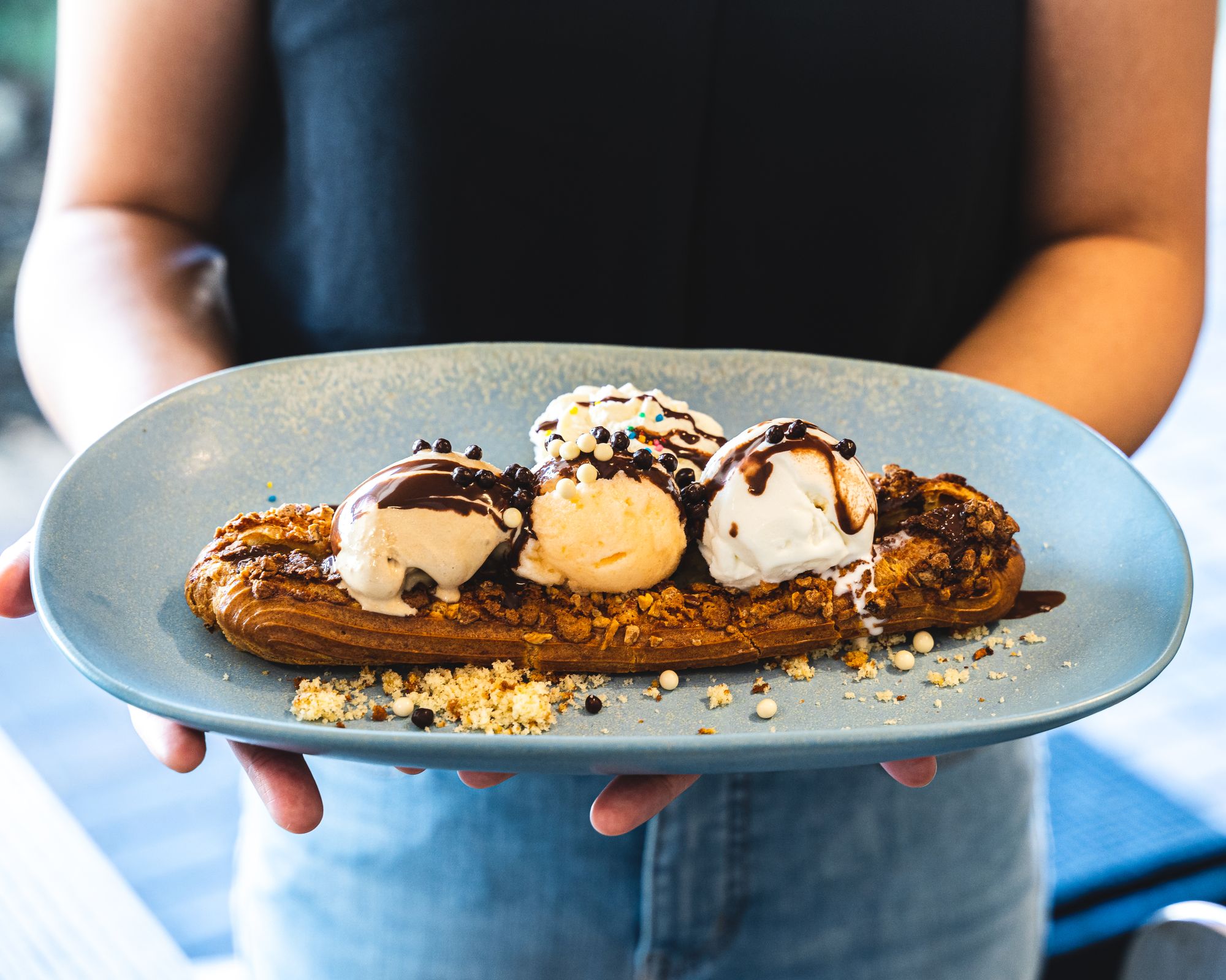 An eclair looking pastry with three scoops of ice cream with spinkles and chocolate sauce, held on a plate