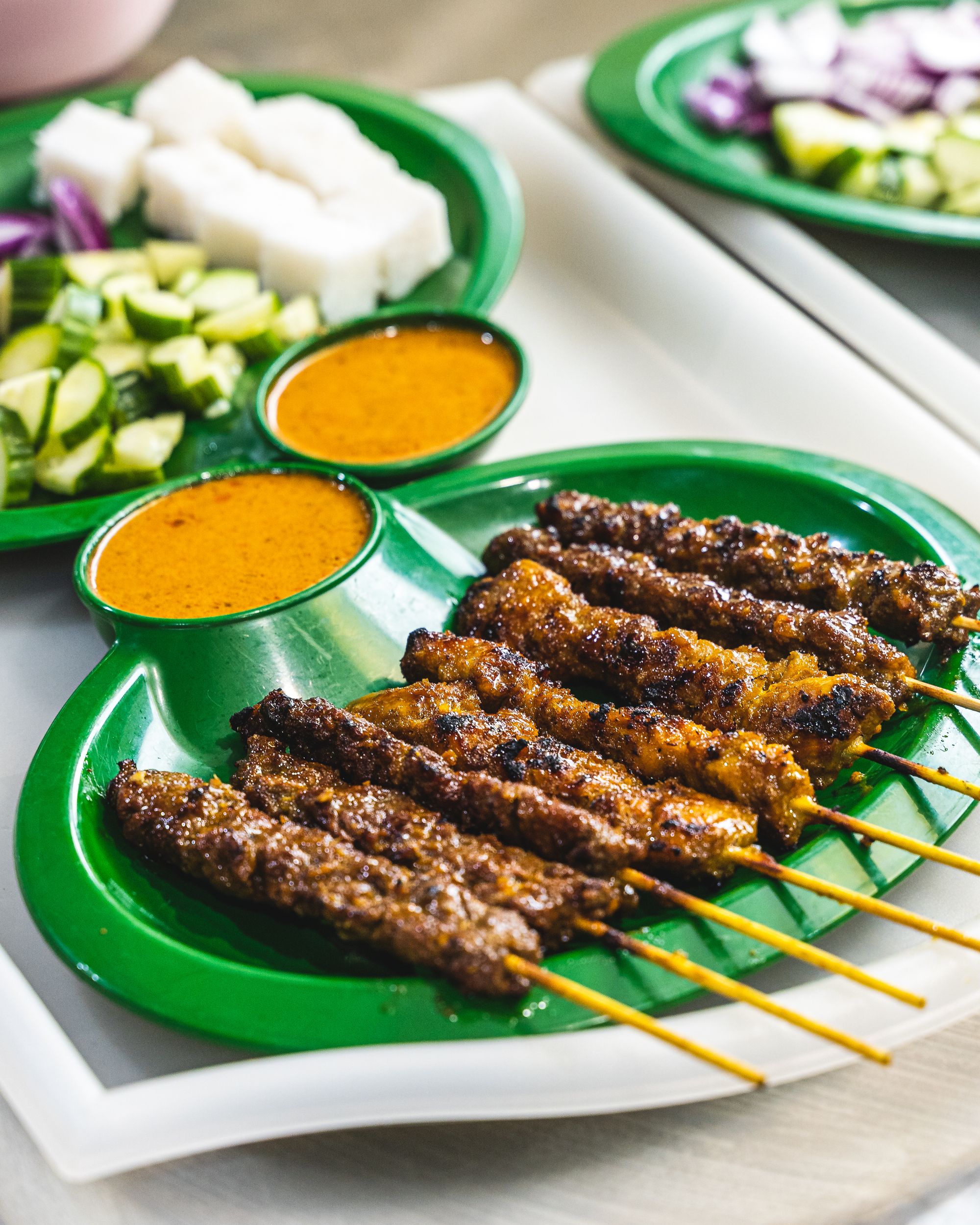 A close up shot of 8 satay sticks, satay sauce and cucumber and rice in the background