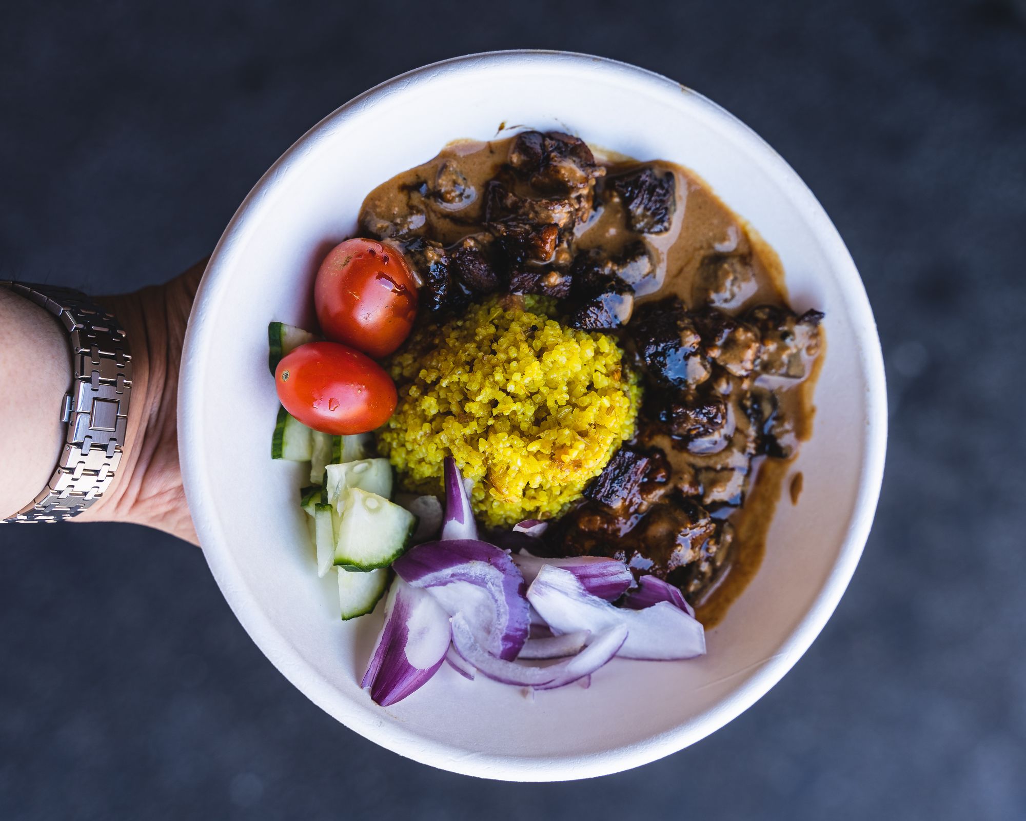 Overhead shot of hand holding a bowl with yellow rice, satay sauce on lamb and assorted vegetables