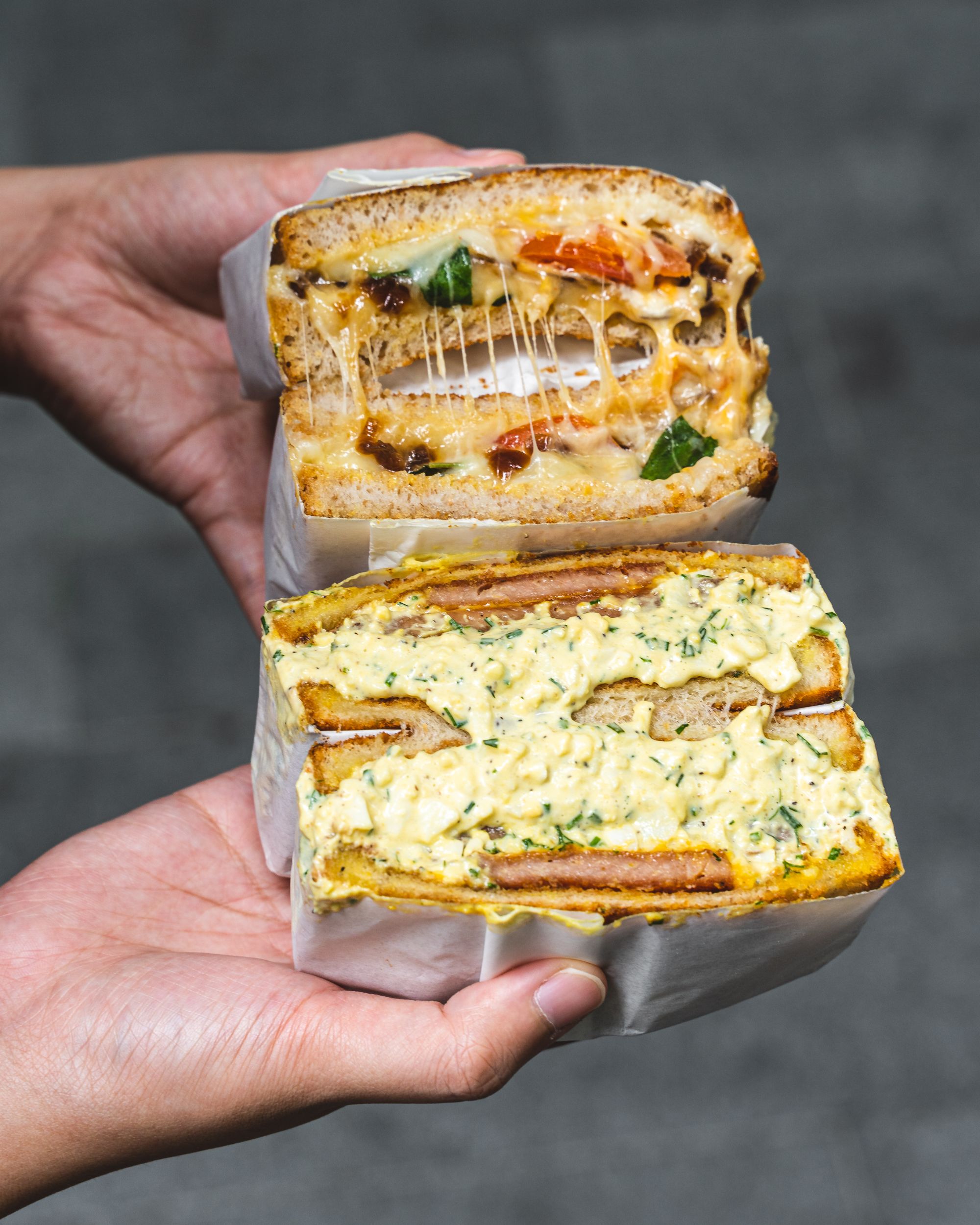 Close up shot of hands holding two sandwhiches cut open, showing the oozing ingredients