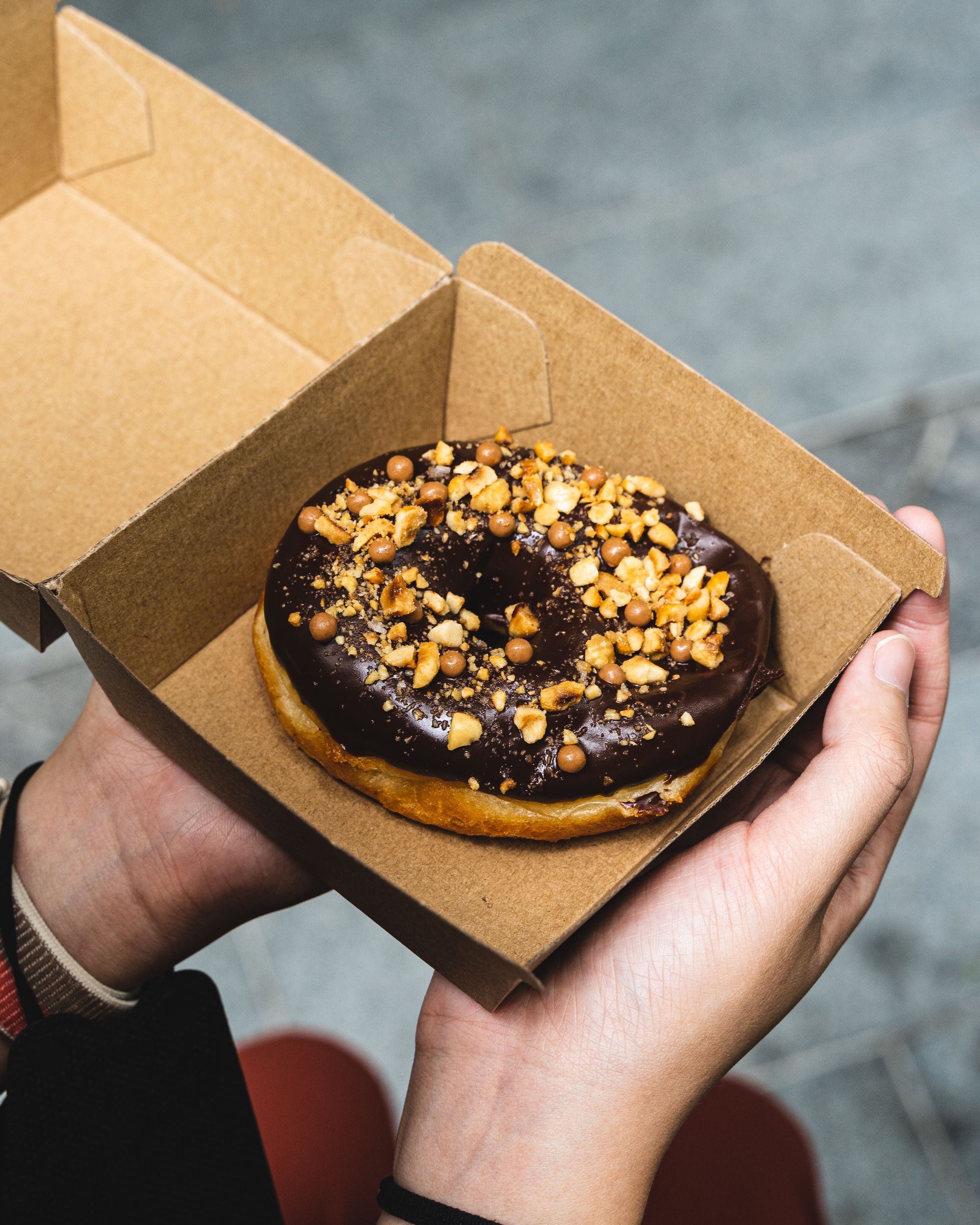 Close up shot of a hand holding a milk chocolate doughnut with crushed hazelnut on the top, in a cardboard box