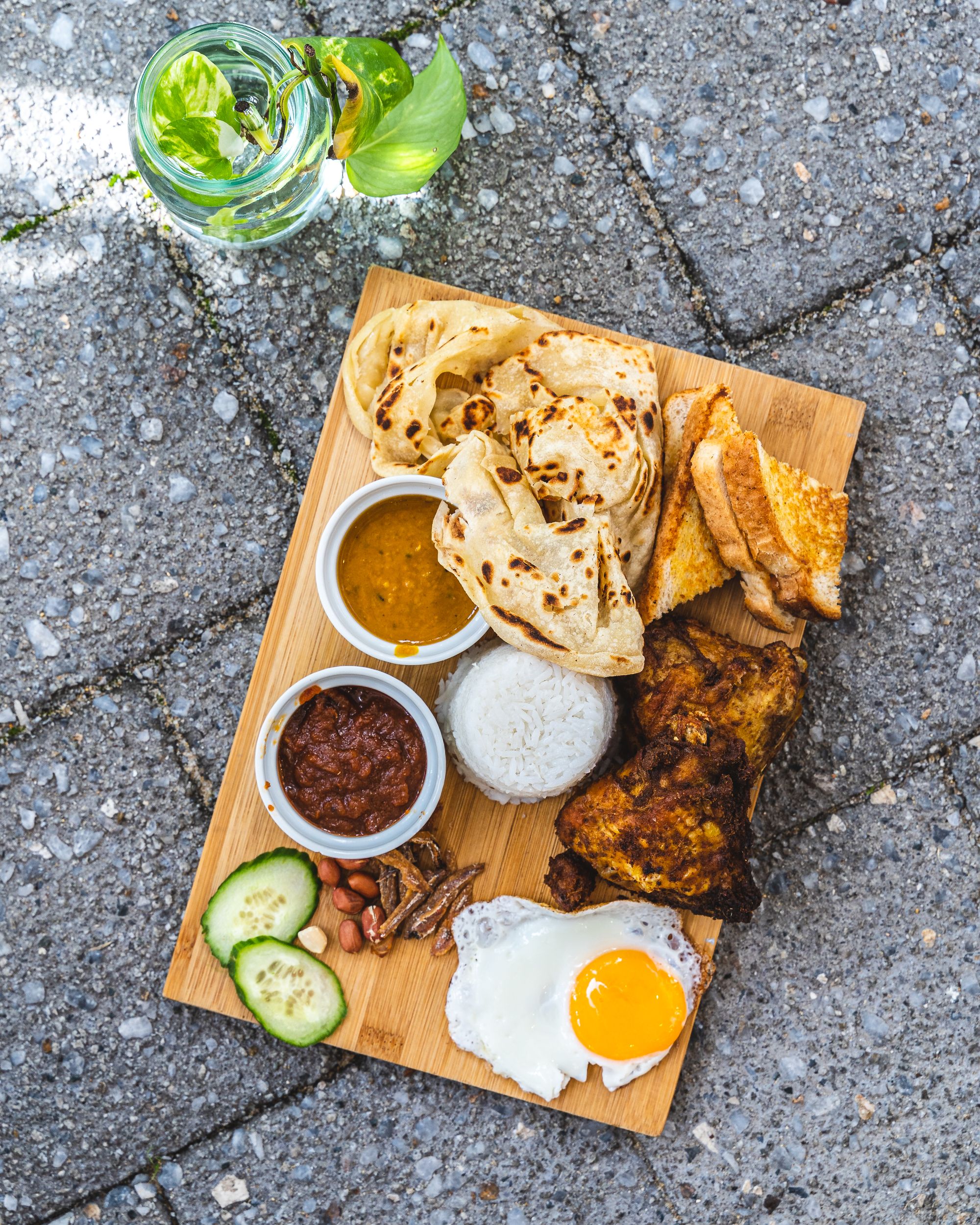 Chopping board on a brick floor with a plant to the right, filled with egg, fried chicken, toast, roti, rice and condiments in ramikins