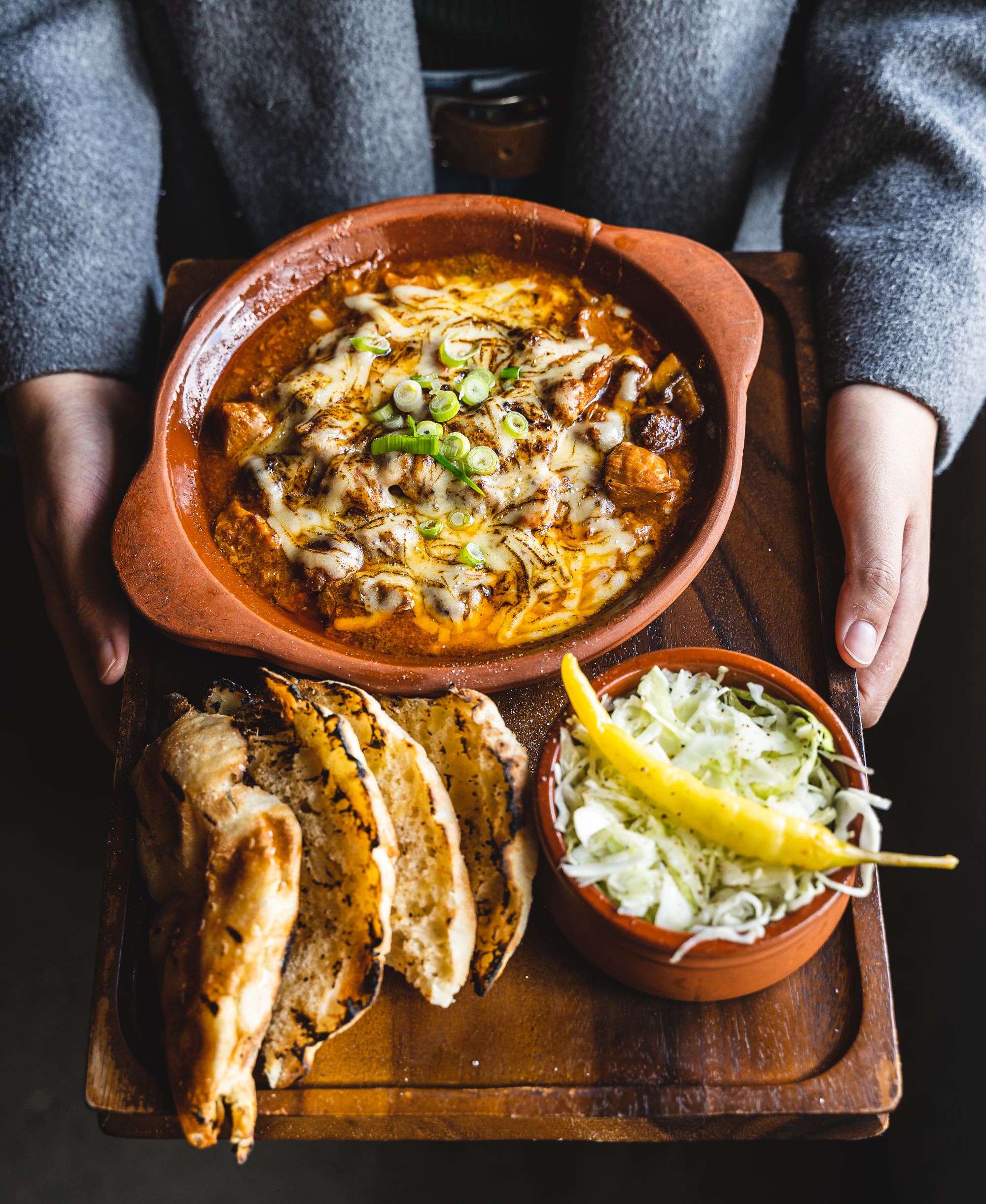 Hand holding a board with a claypot dish that has cheese ontop of meat, turkish bread and a bowl of cabbage with a yellow chilli on-top