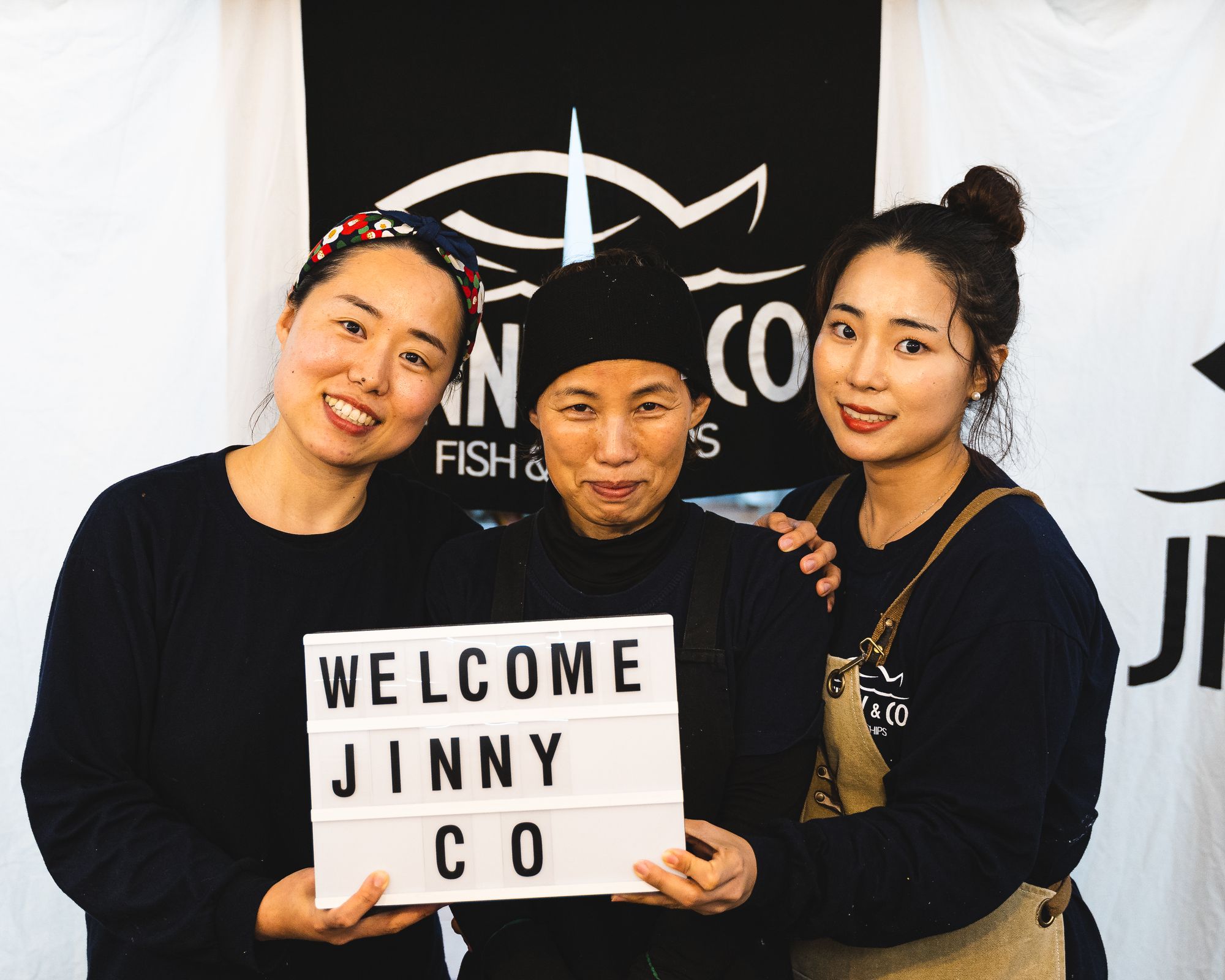 Three Asian women holding a sign that says "Welcome Jinny Co"