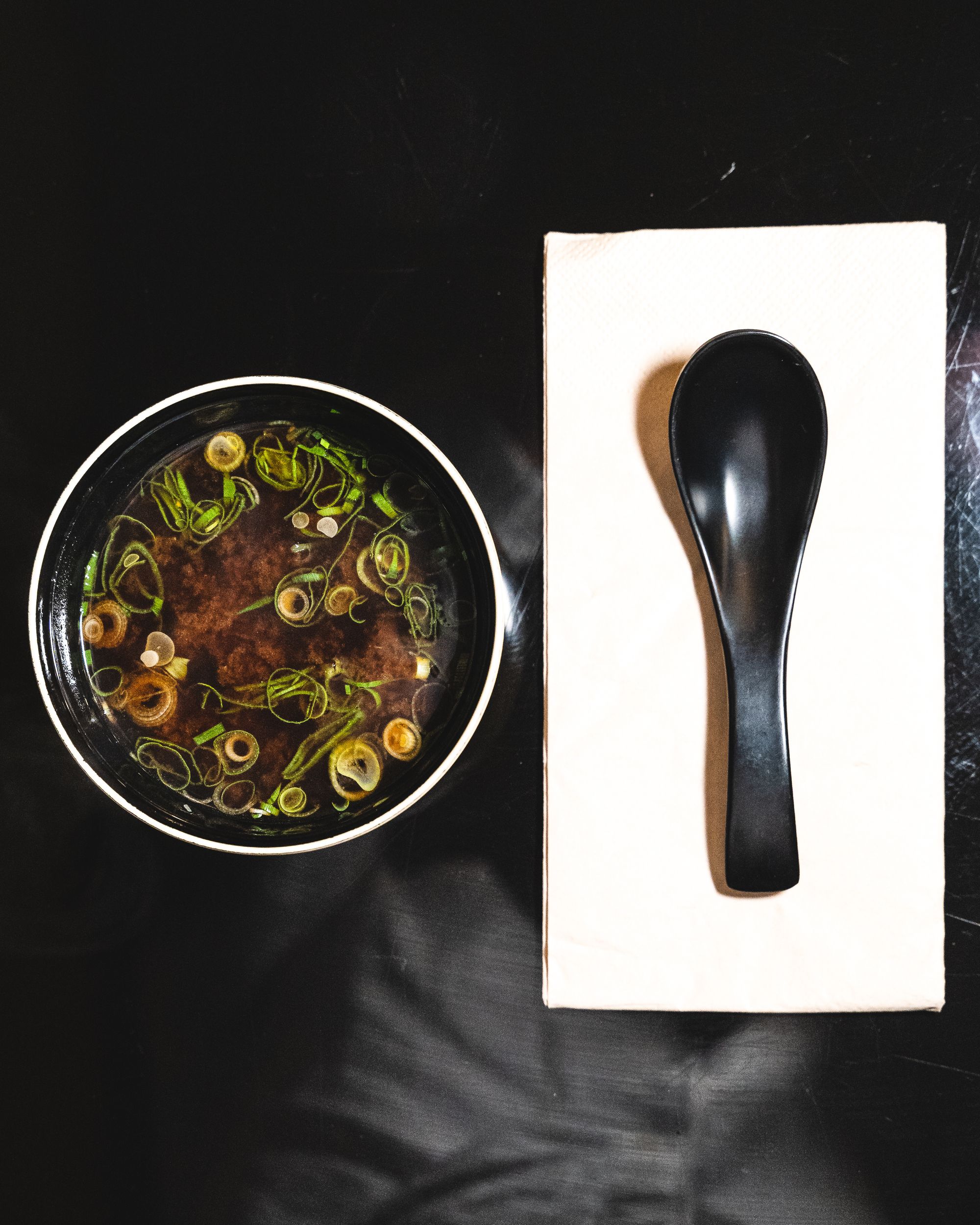 A bowl of miso soup next to a soup spoon sitting ontop of a white napkin