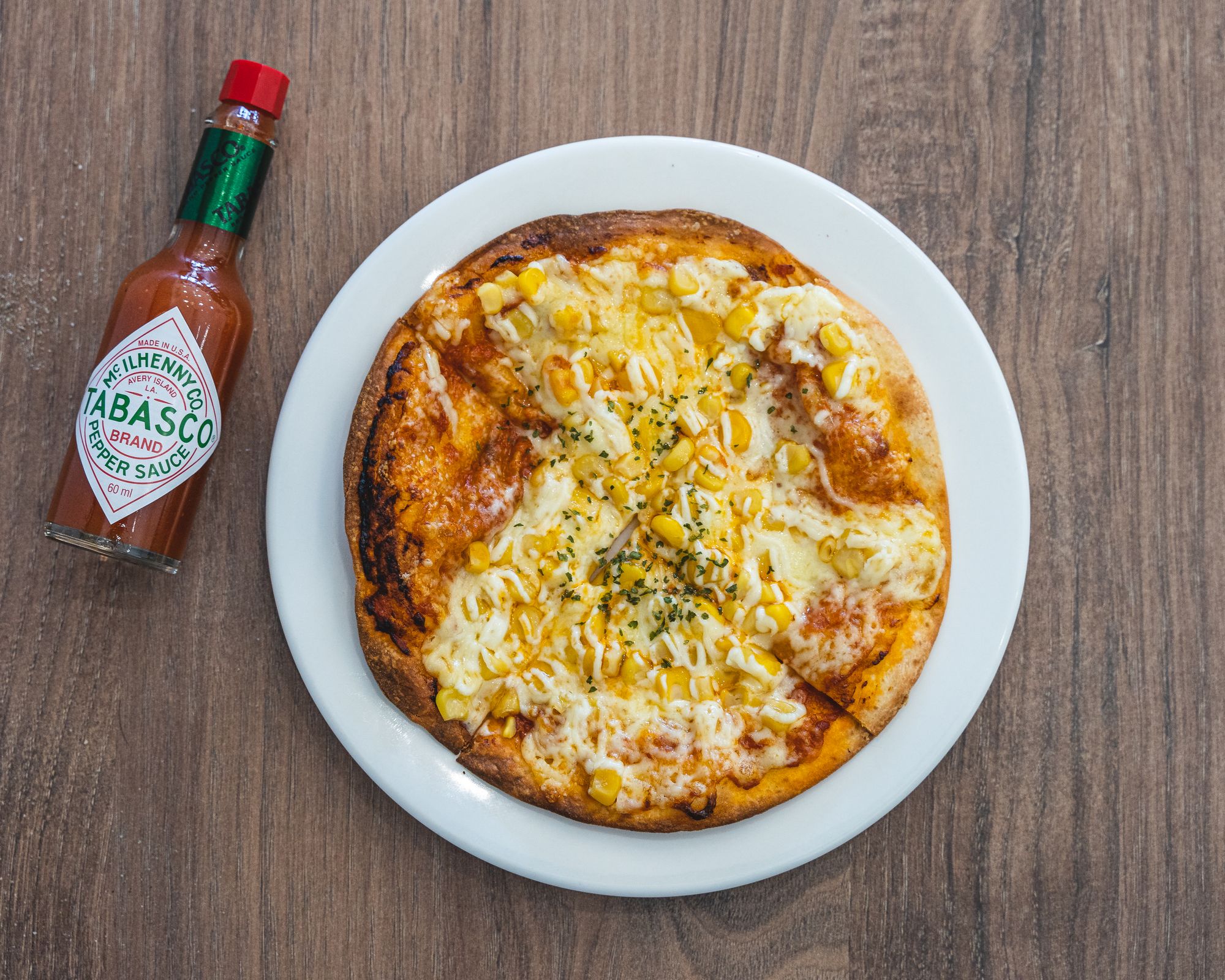 Overhead shot of tabasco sauce next to a corn and mayonaise pizza on a white plate