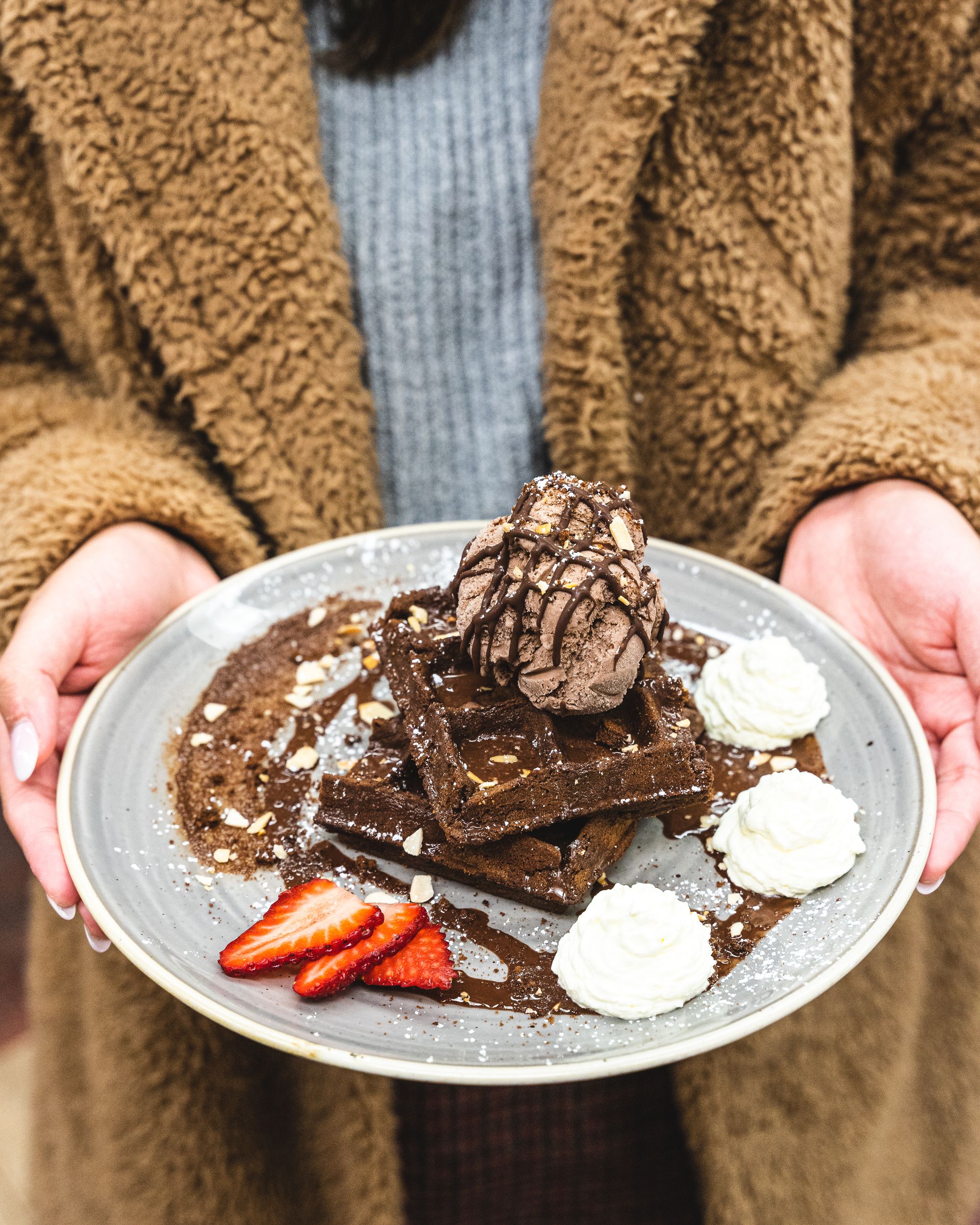 Pedicured hands holding a plate with chocolate waffles with a scoop of chocolate ice-cream on top