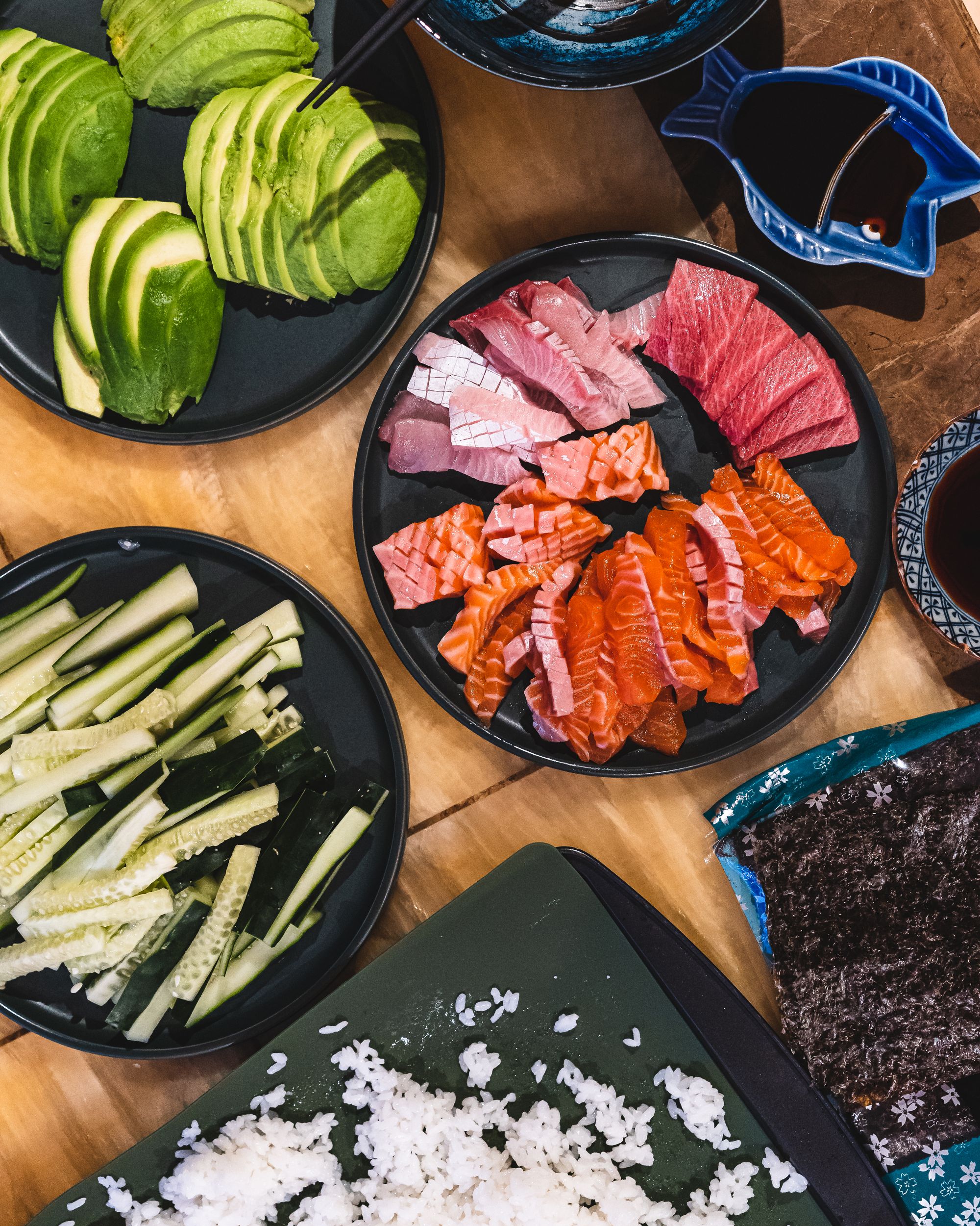 A flat lay showing avocado, sashimi, rice and cucumber on seperate plates