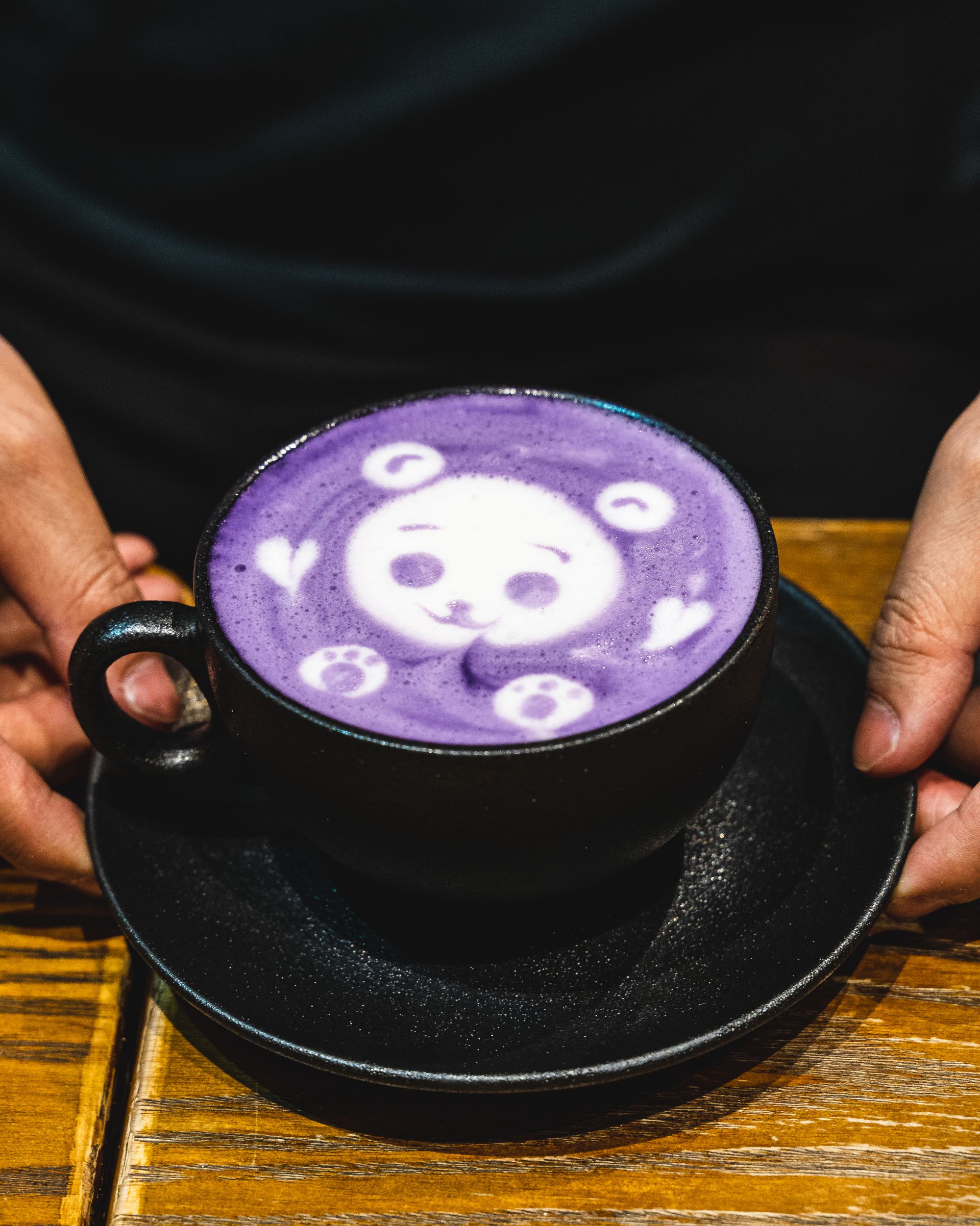 Hands holding a black mug with a purple coloured latte, topped with a teddy bear for latte art