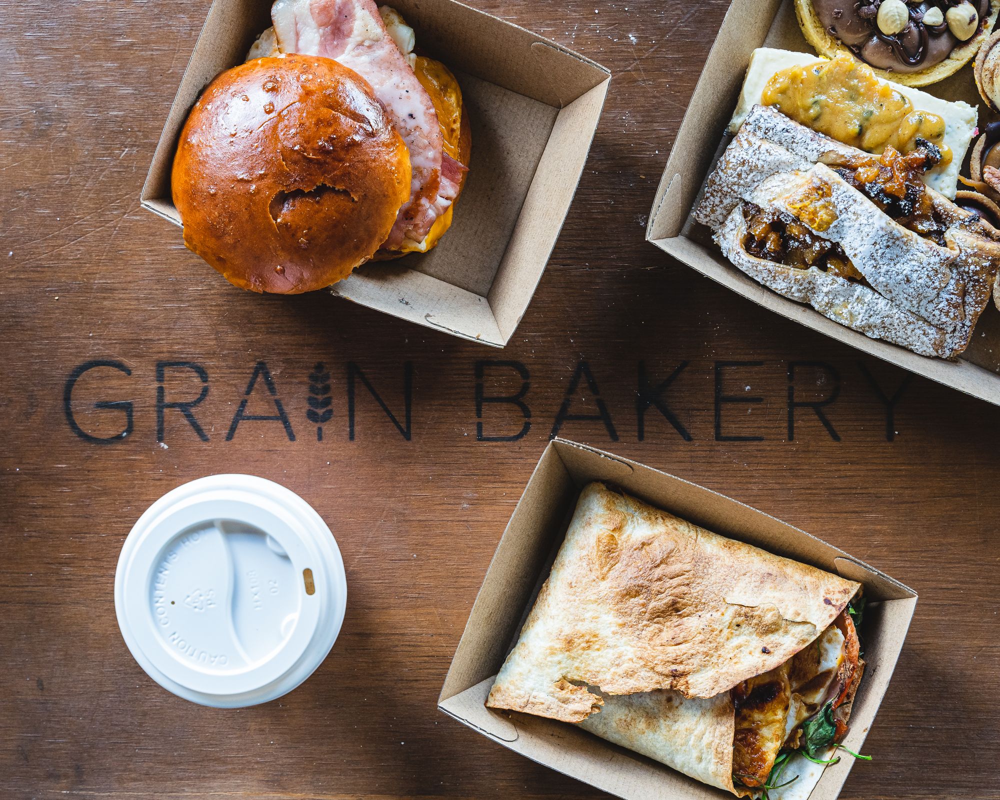 Flat lay with a burger, wrap and pastries on-top of a wooden board that says "Grain Bakery"