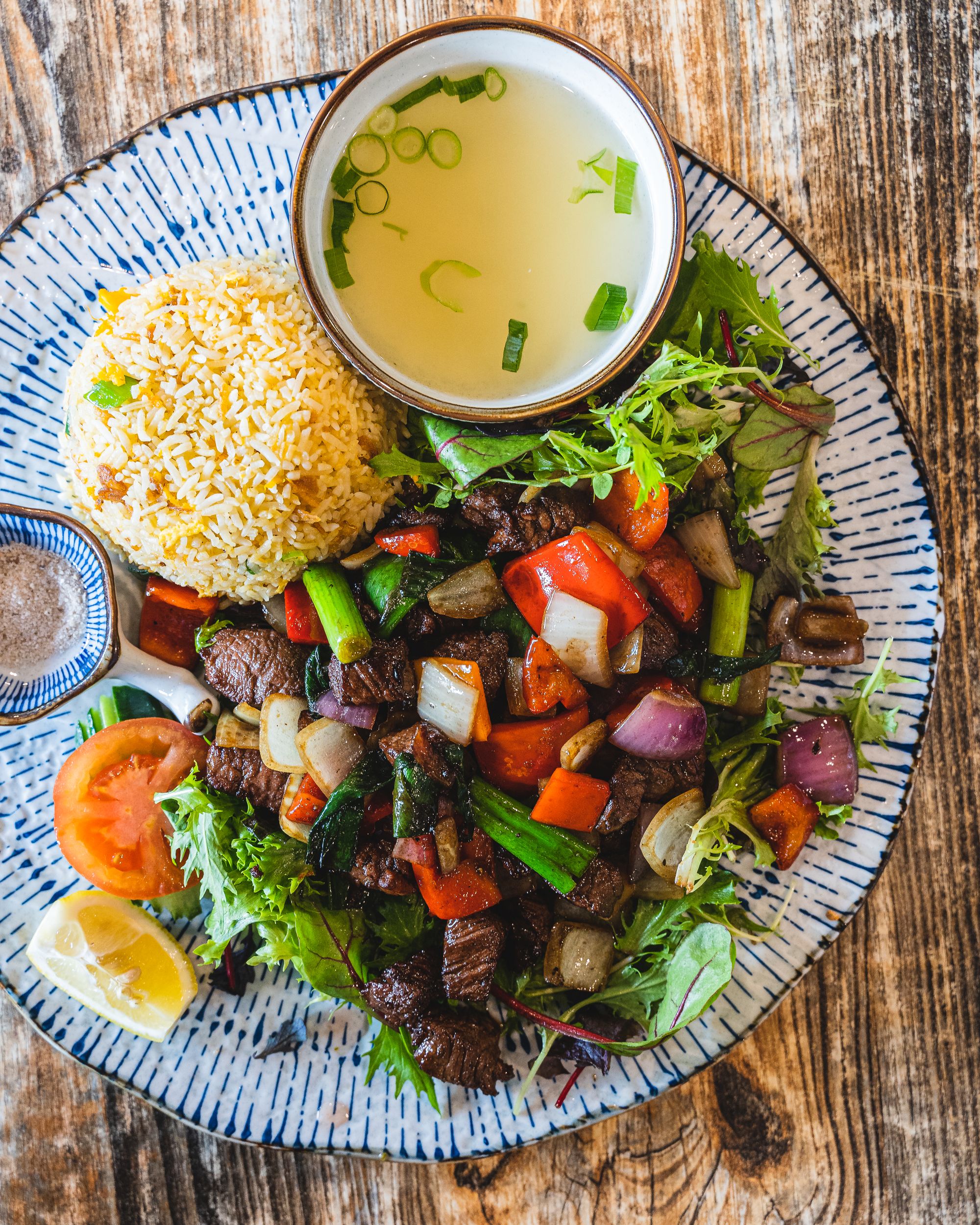 Top down shot of a palte with fried rice, stir fried beef and vegatables, a bowl of soup and a slice of lemon