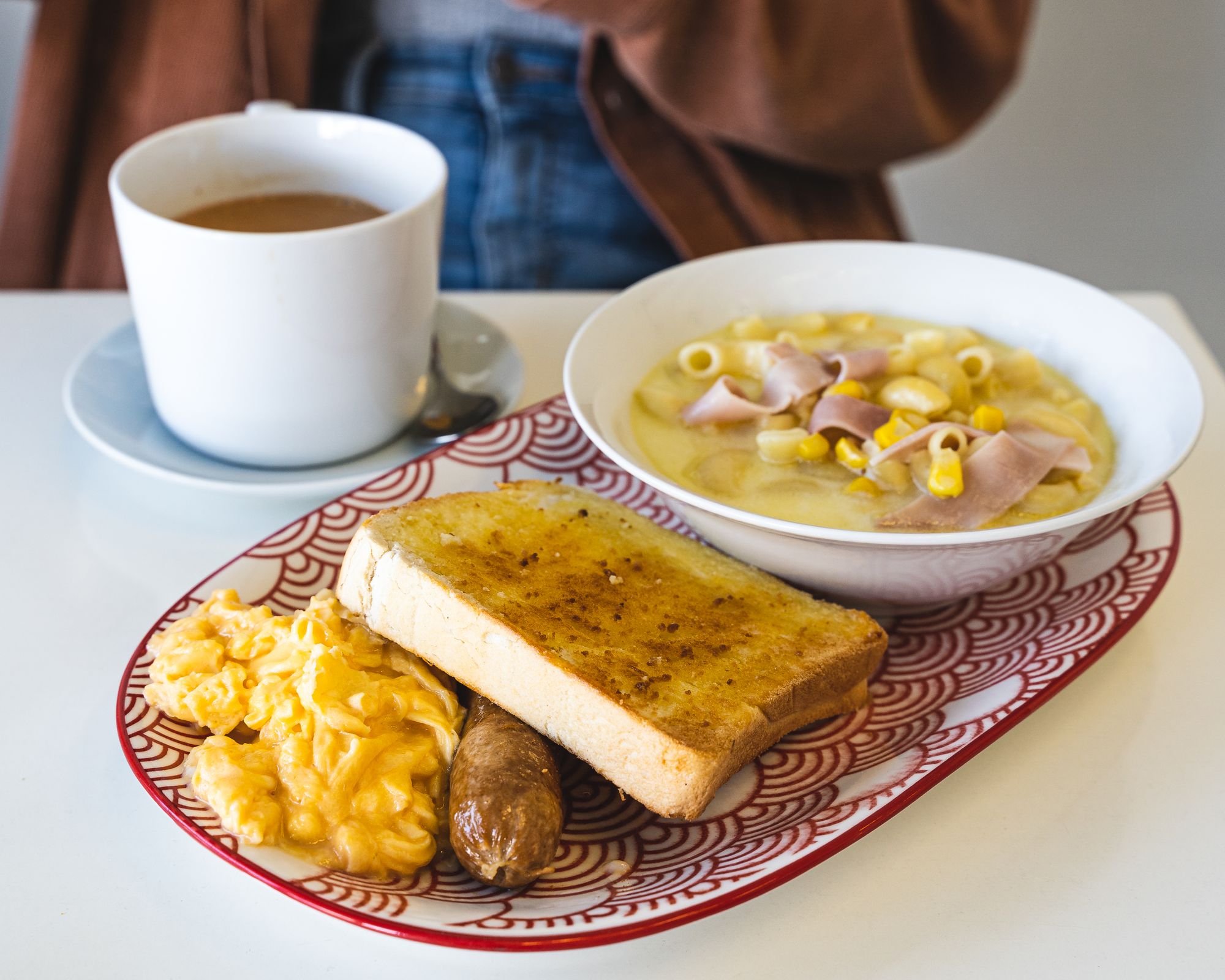 Plate with egg, sausage, toast, a bowl of macaroni and ham soup and a tea cup