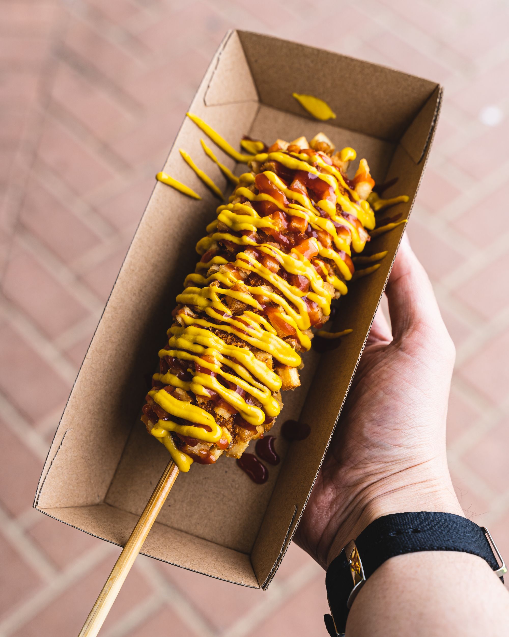 Hand holding a Korean street dog in a tray, covered in mustard and tomato sauce