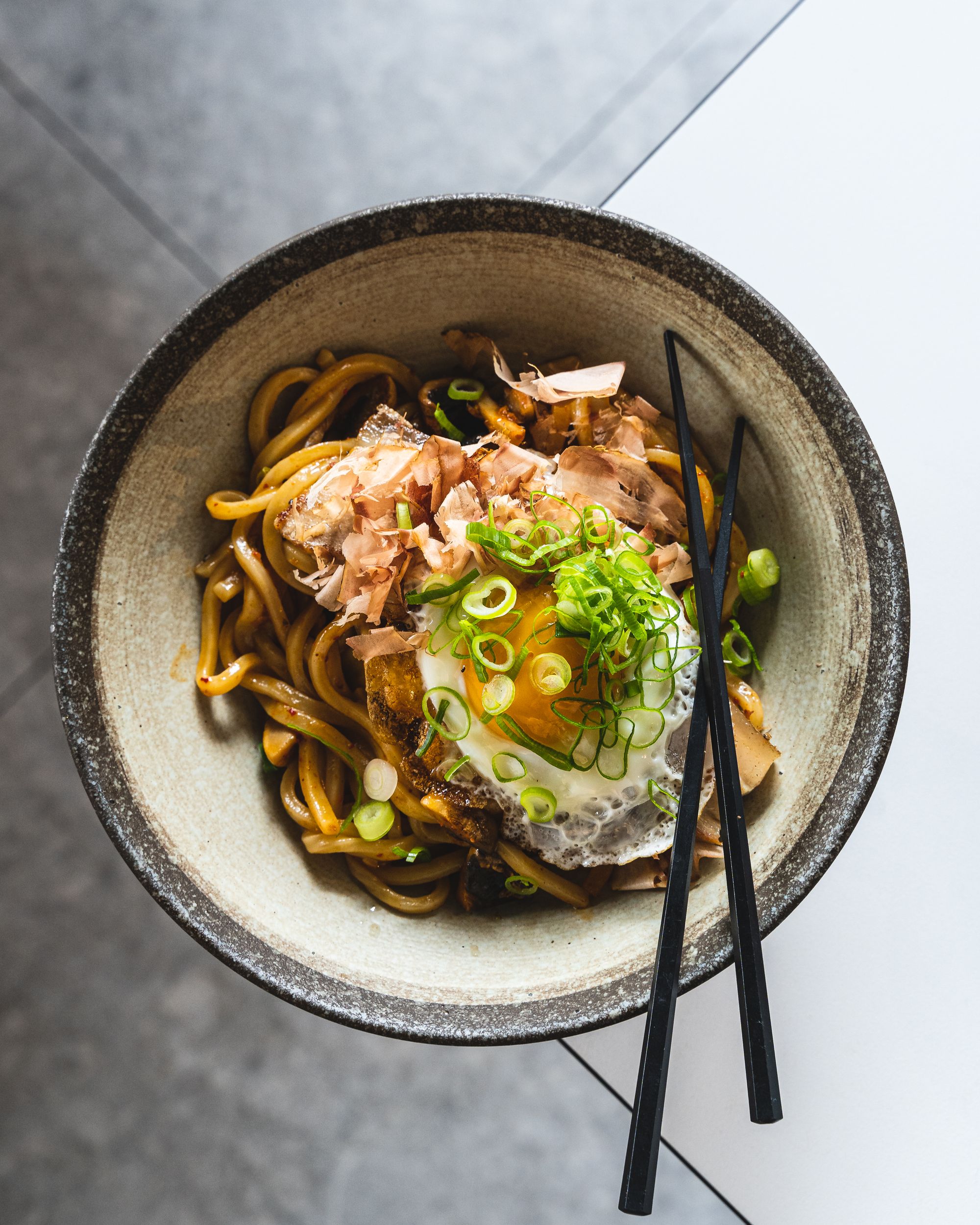 Top down close up of noodles in a bowl with a fried egg, spring onions and a pair of chopsticks