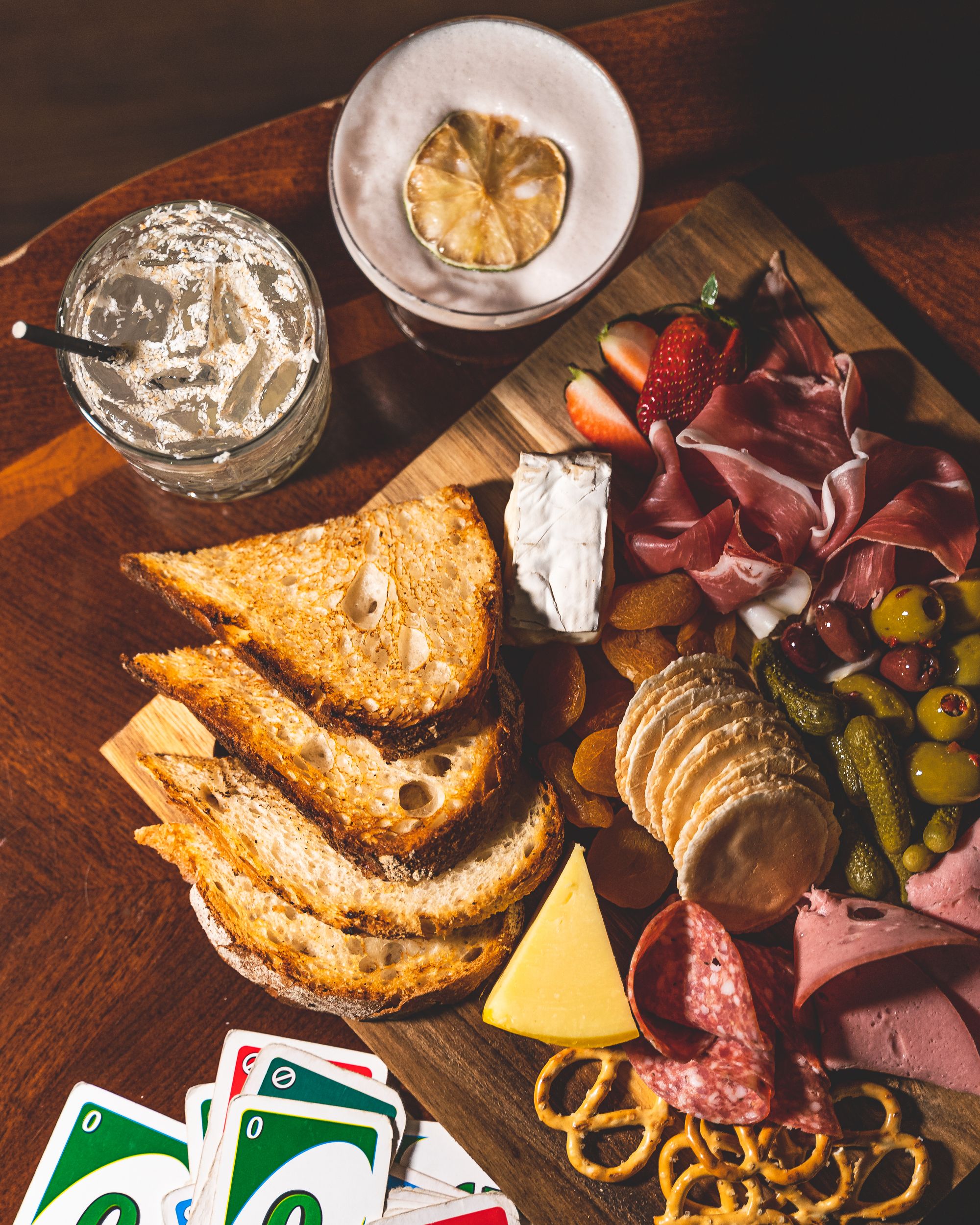 Flat lay of charcuterie board with two cocktails on the top and uno cards in the bottom