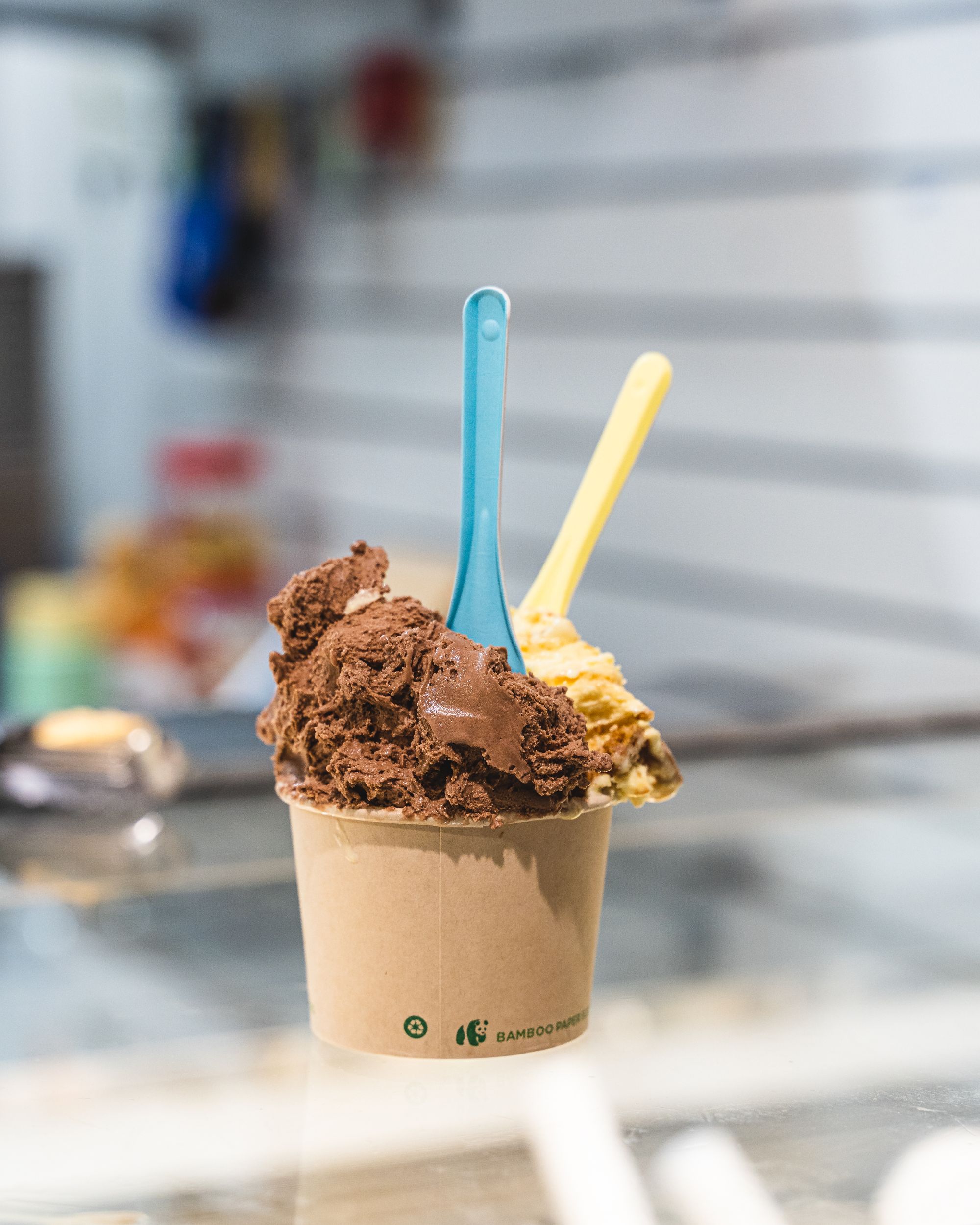 Two scoops of gelato in a paper cup sitting on-top of a gelato fridge