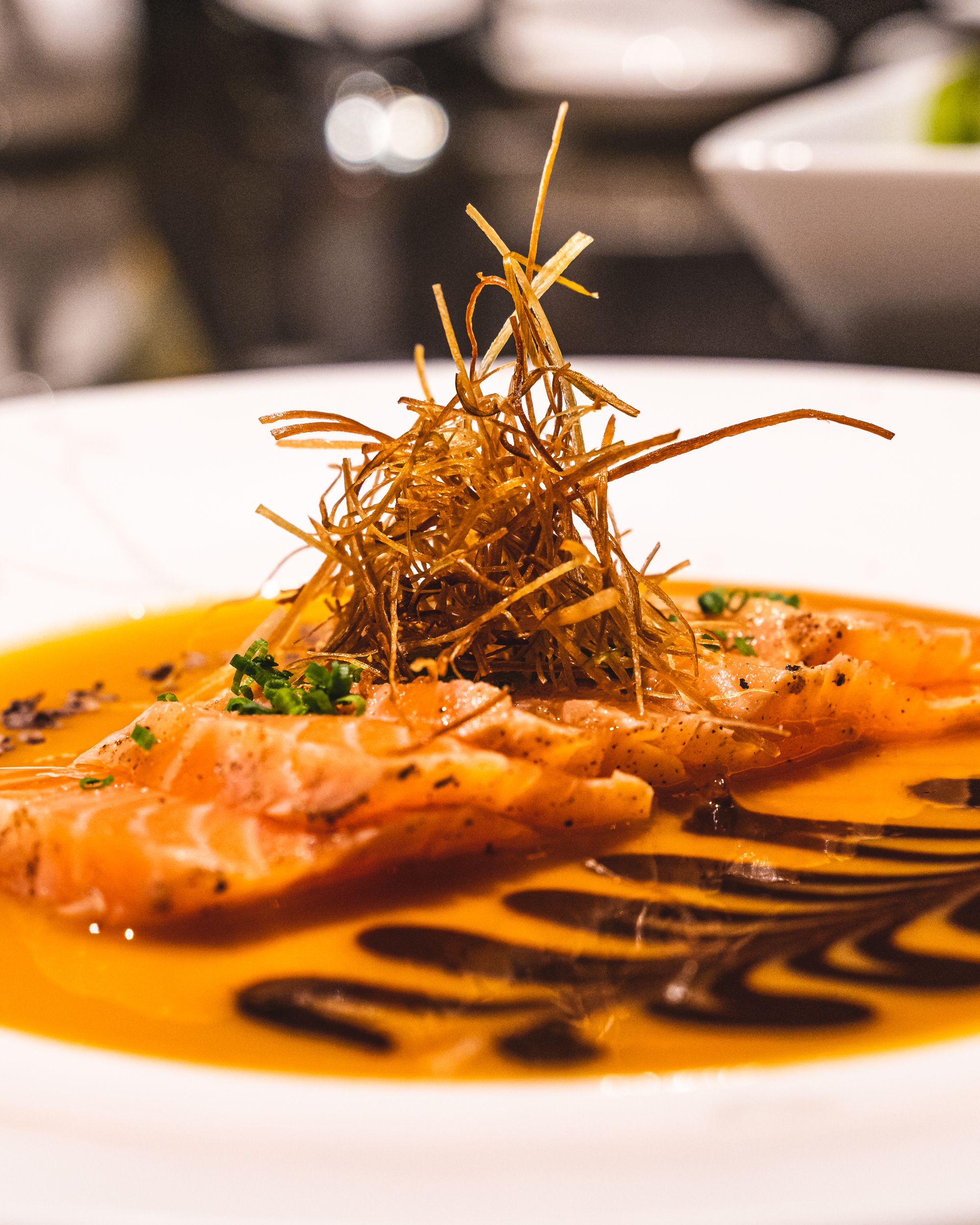 Close up of salmon belly sashimi with crispy leeks piled on top and orange coloured sauce