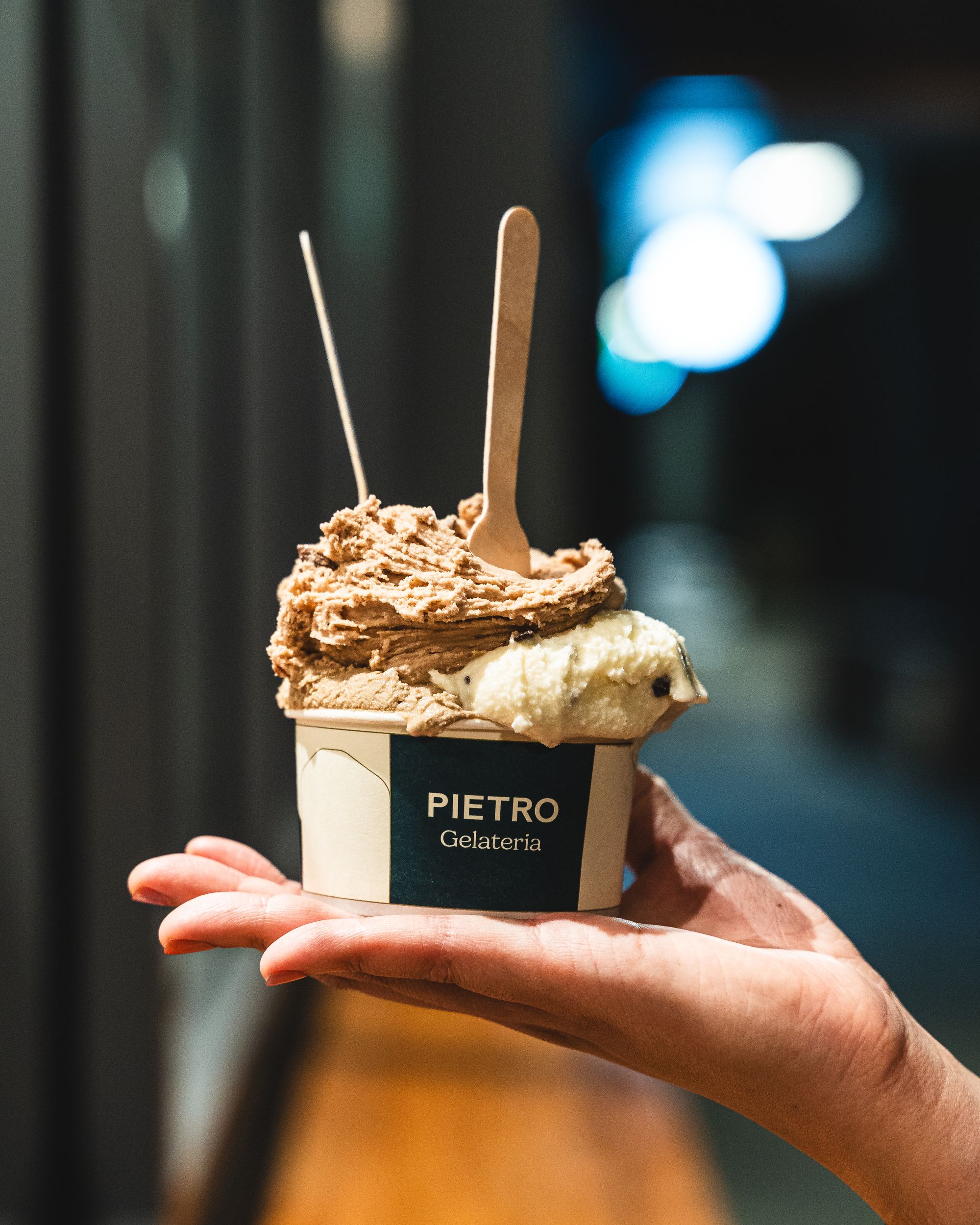 Hand holding a Pietro Gelateria branded gelato cup with gelato and spoons sticking out of it 