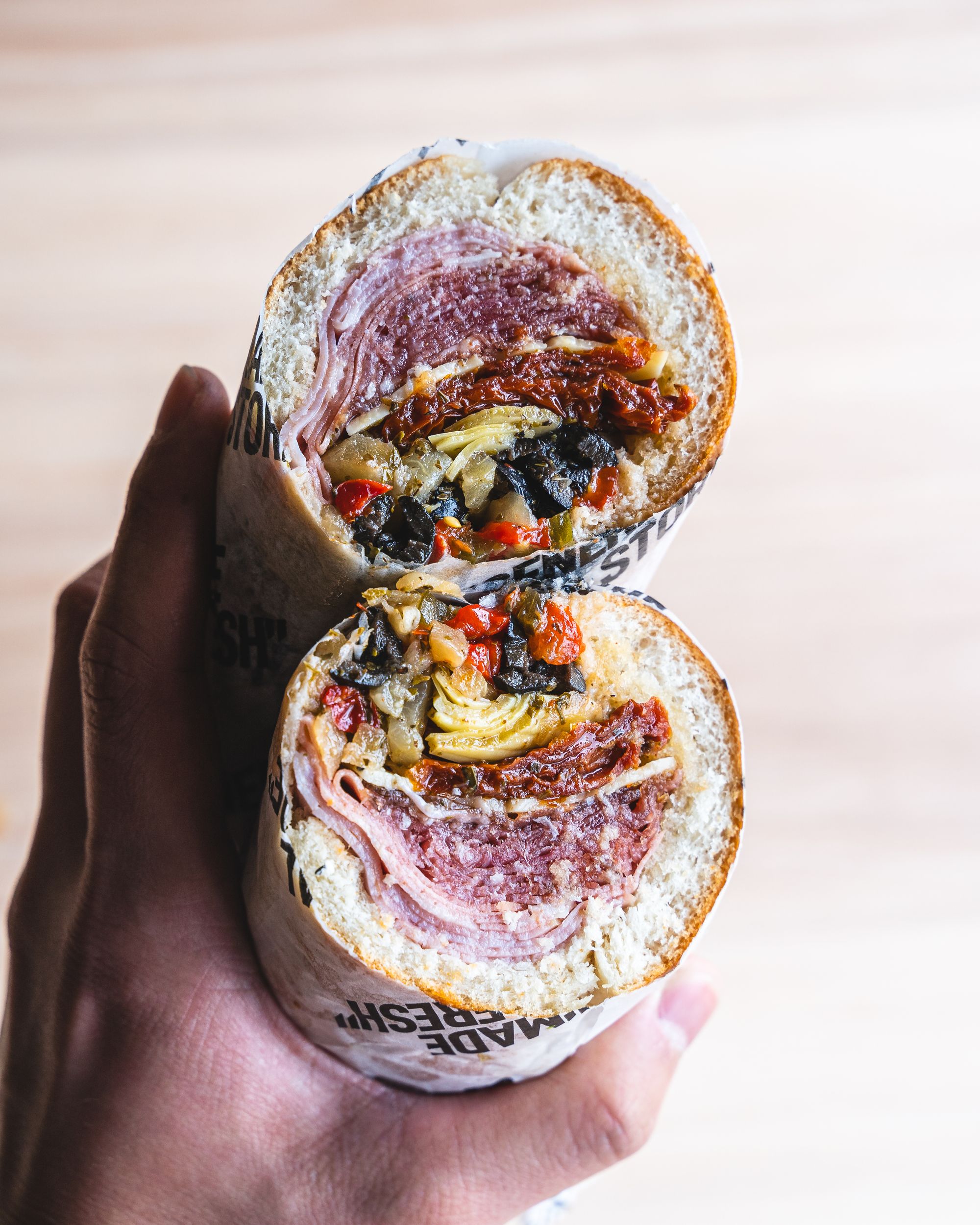Hand holding two sandwich rolls showing a cross section of Italian cured meats and antipasto
