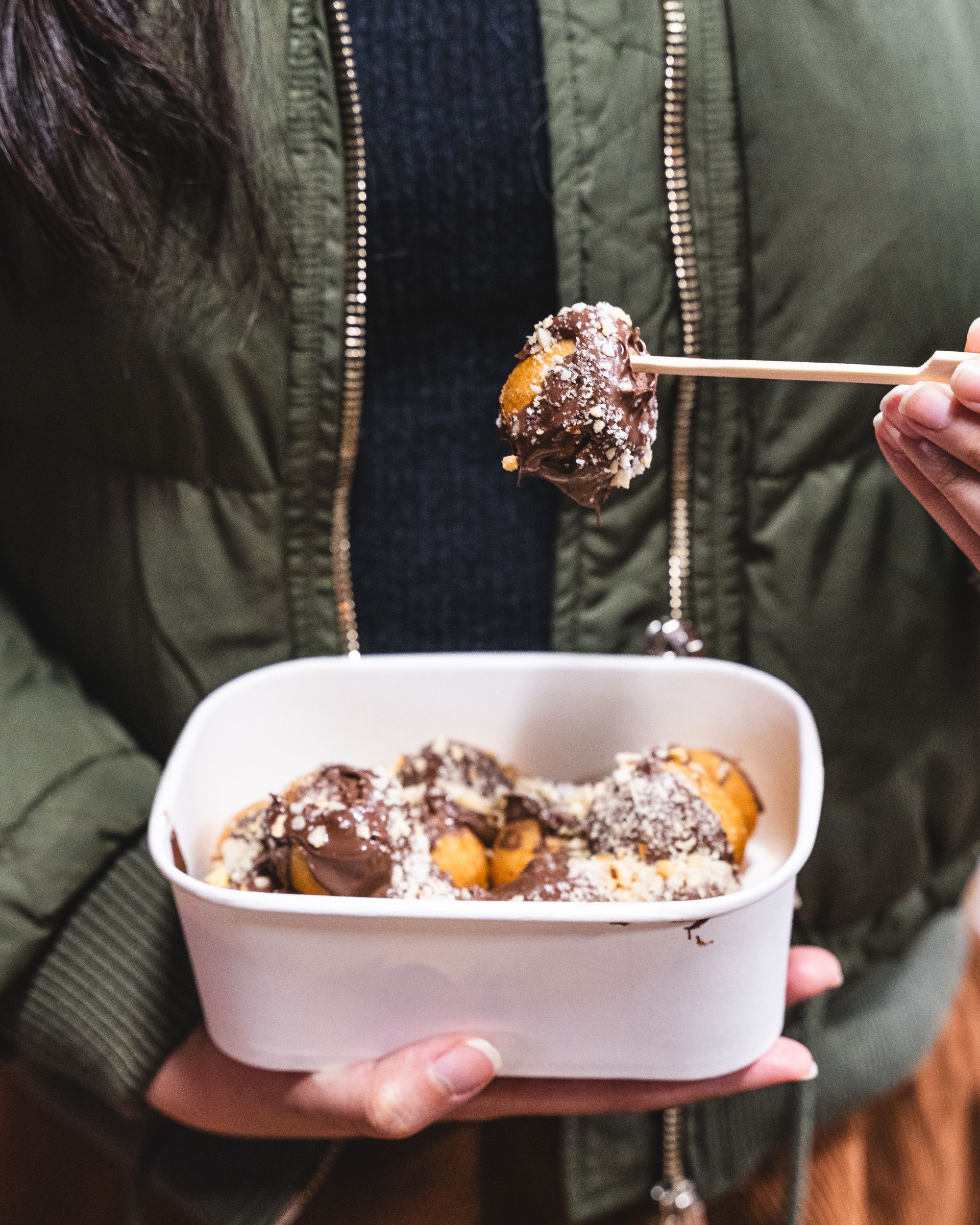 Hand holding a paper container of fried donut balls topped with Nutella