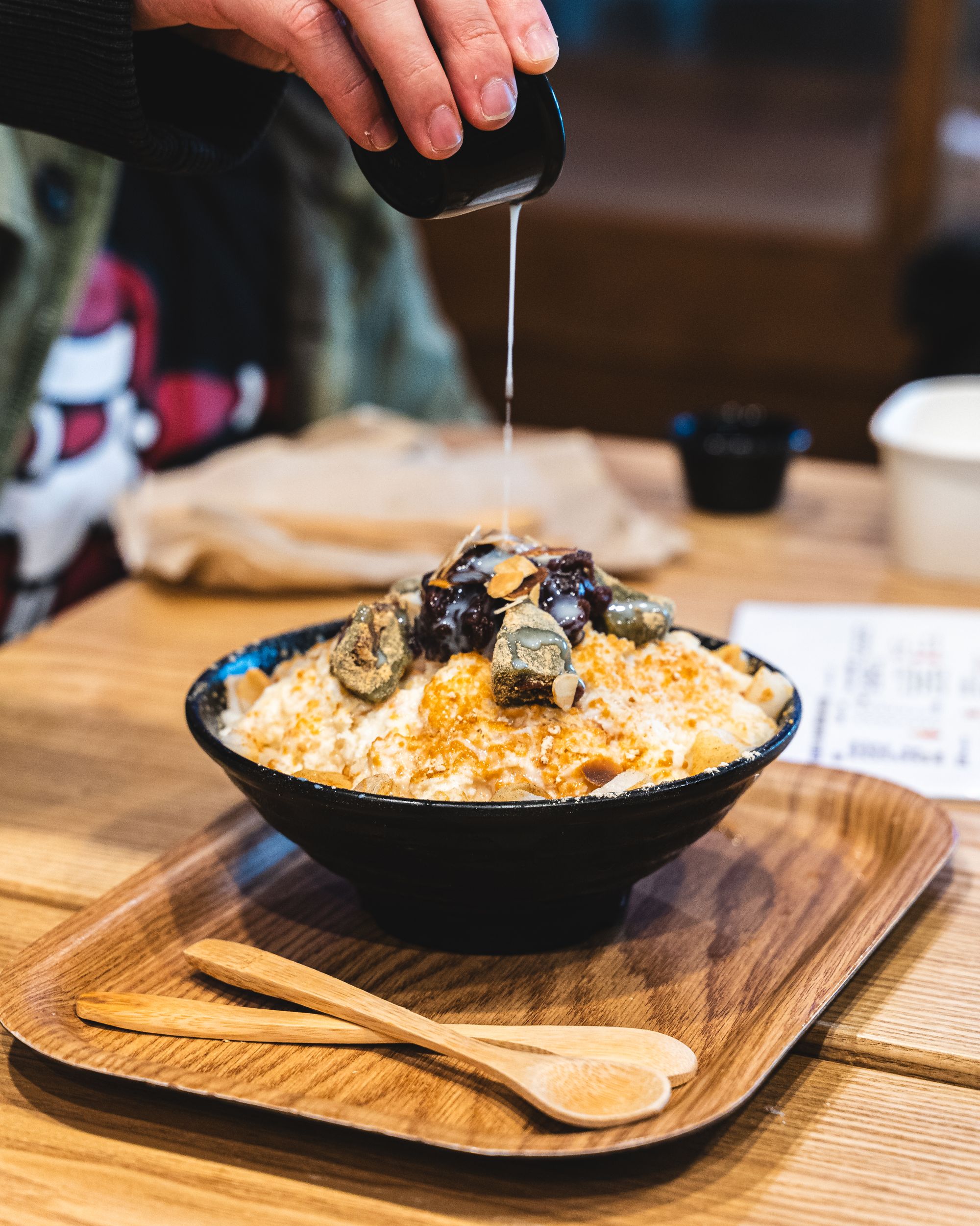 Hand pouring condensed milk onto bingsu which is topped with red bean and mochi and roasted soy powder