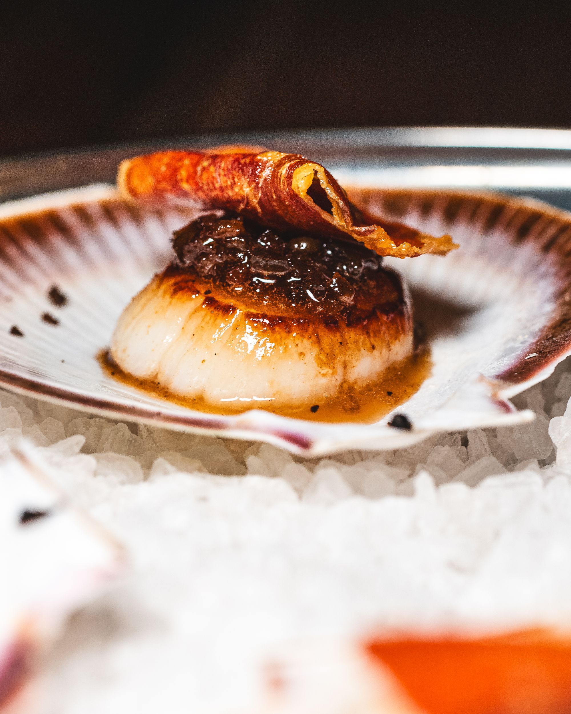 Close up of scallop topped with bacon, sitting on-top of a scallop shell