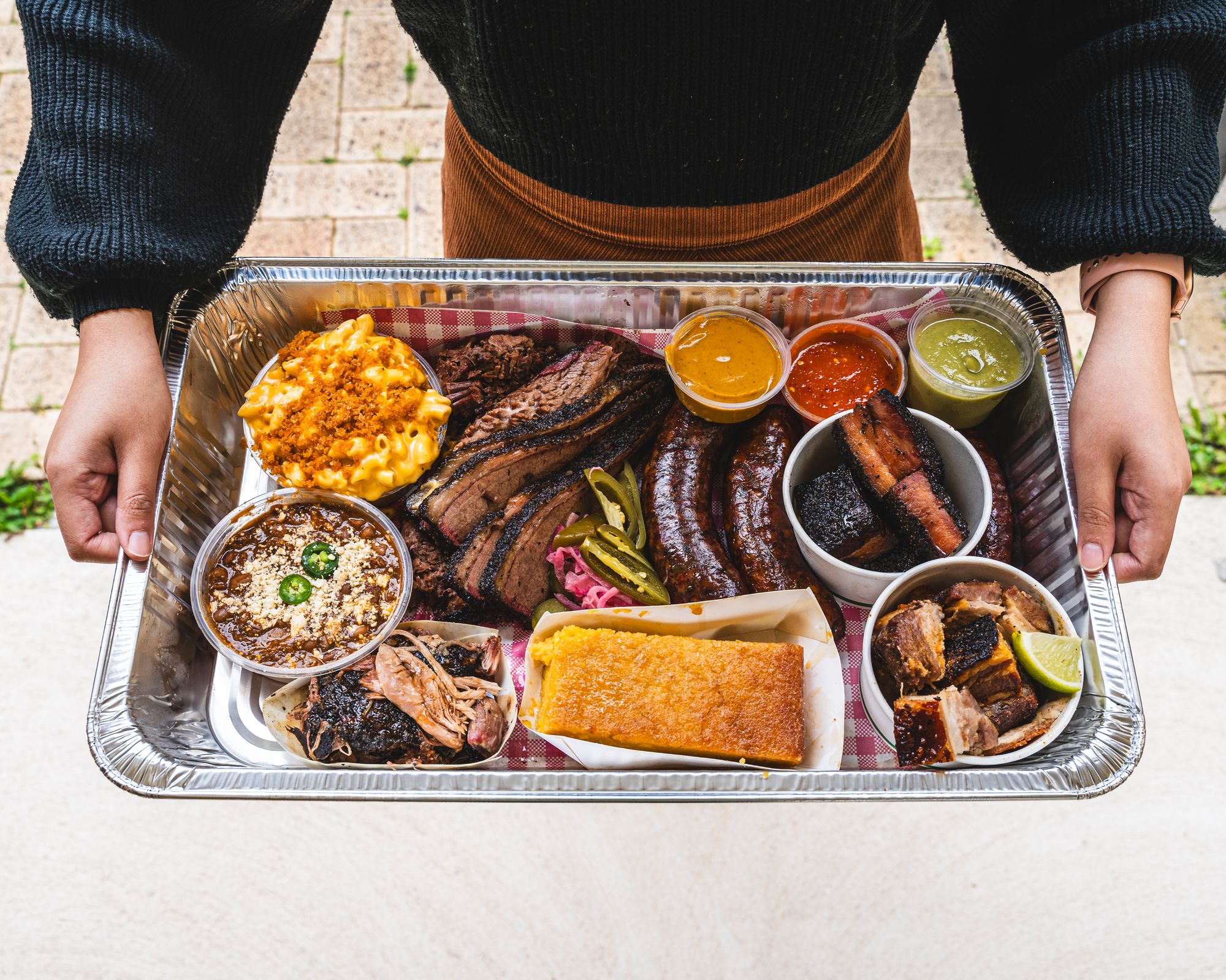 Hand holding metal tray with assorted American BBQ meat, cornbread, mac and cheese, chicharron and sauces