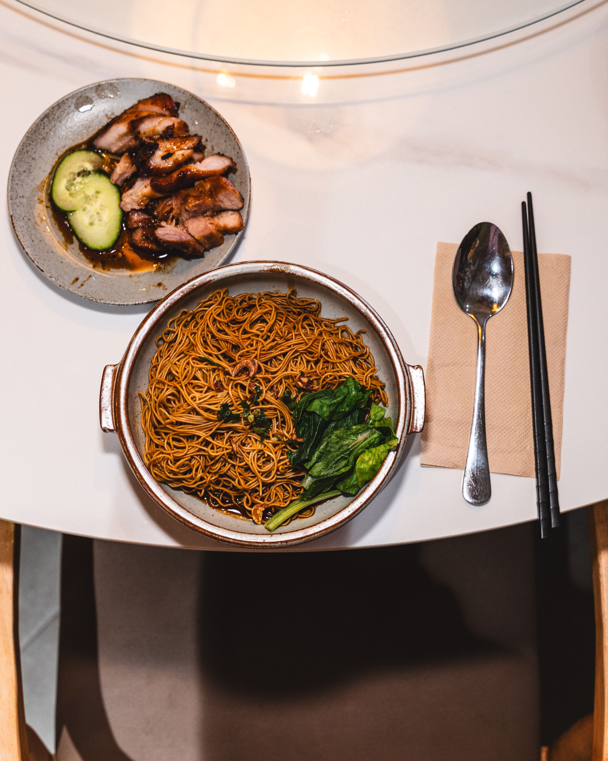 Top down shot of a plate of noodles in dark soy sauce with Chinese vegetables and a plate of char siew pork