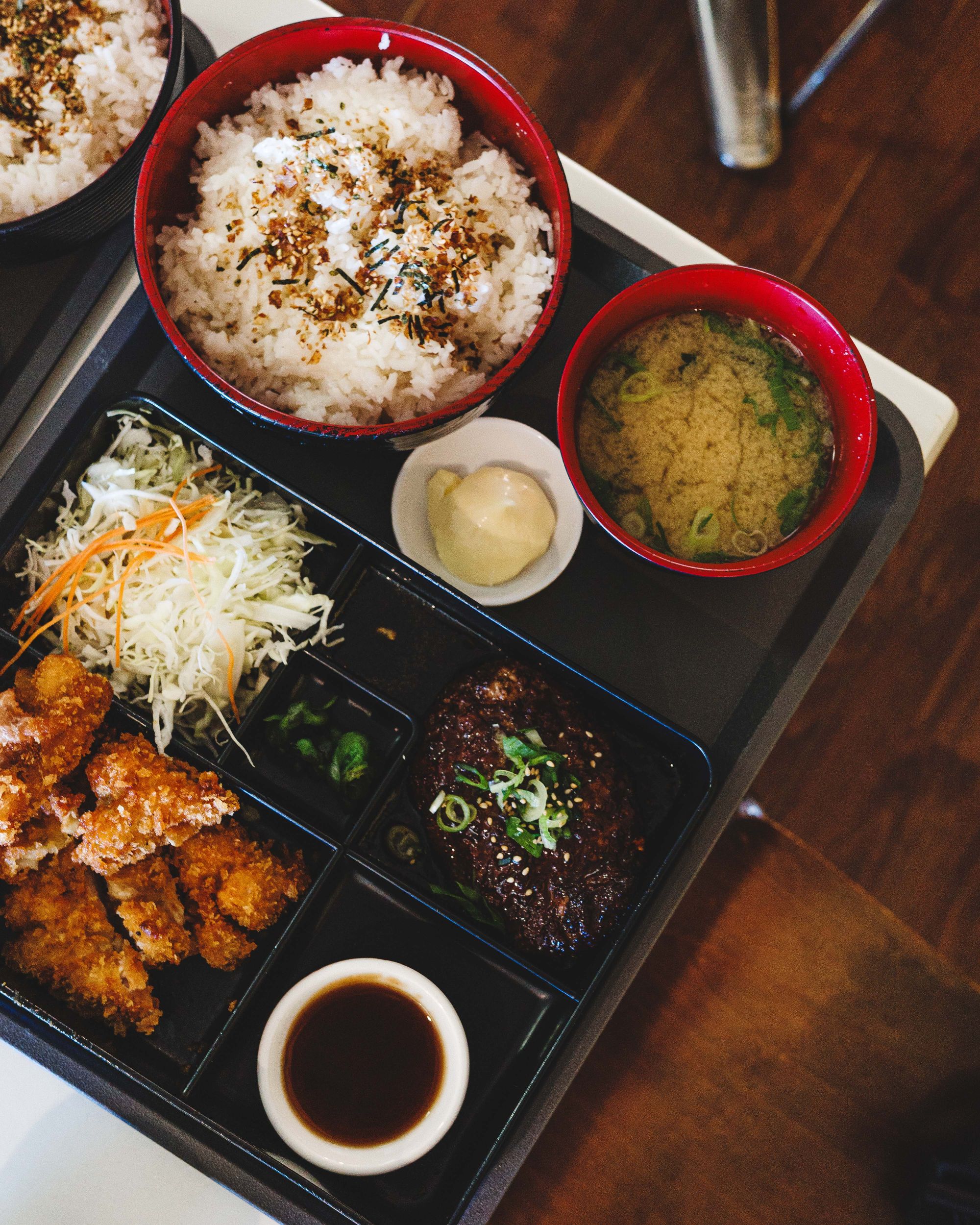 Top down shot of bento with Hamburg steak, chicken katsu, salad, katsu sauce, rice, miso soup and mayonnaise