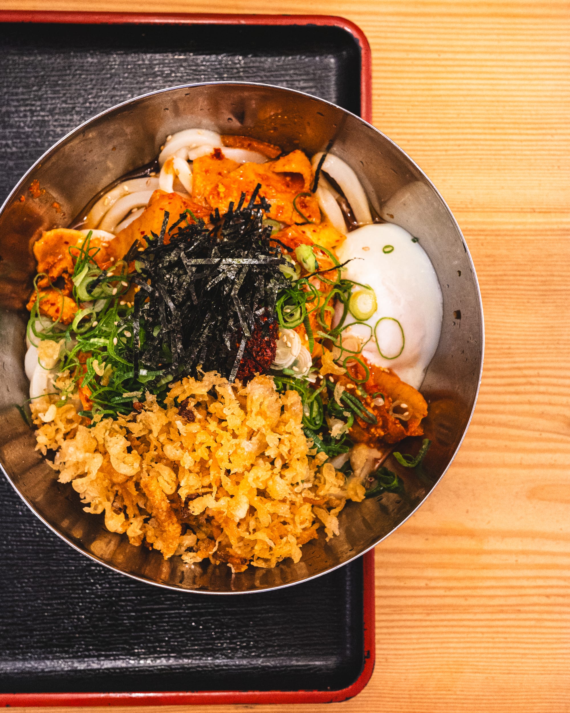 Top down shot of udon in metal bowl with panko, seaweed, onsen egg, kimchi and spring onions