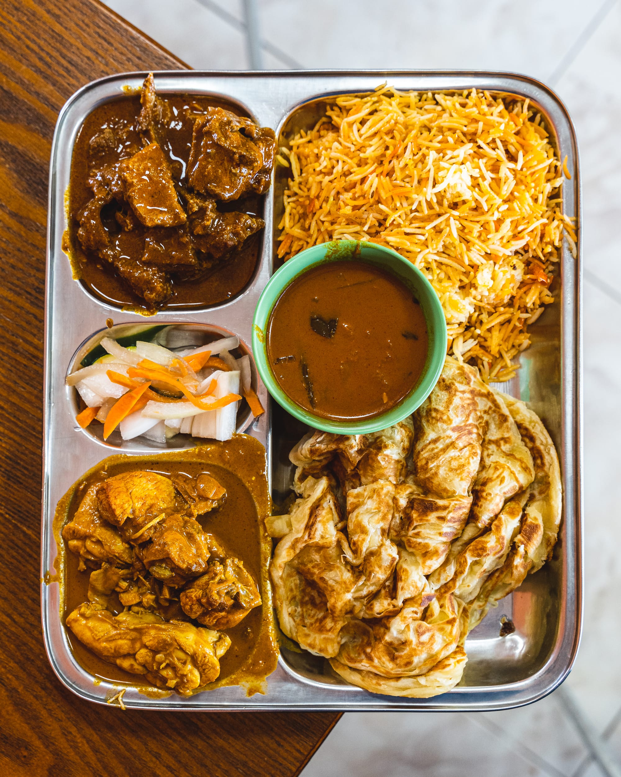 Top down of curry on a silver plate with prata, rice, pickled vegetables and two meat curries