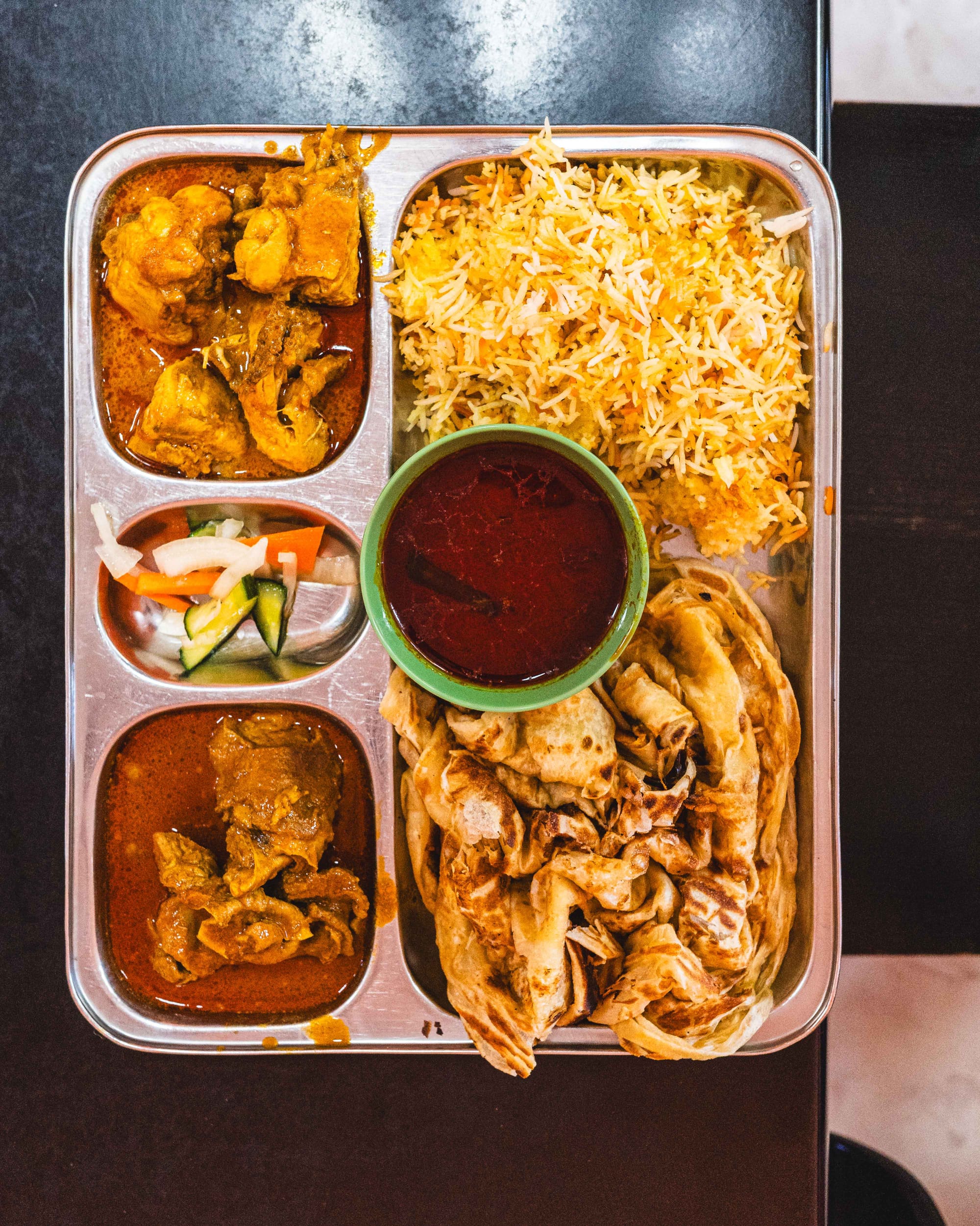 Top down shot of steel tray with curry, pickled vegetables, biriyani and prata