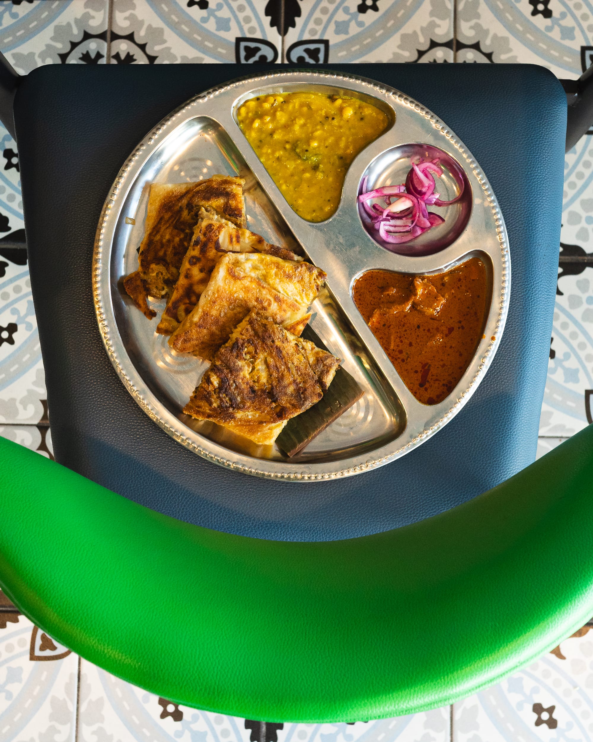 Top down shot of murtabak on a stainless steel tray with dhal, curry and pickled onion