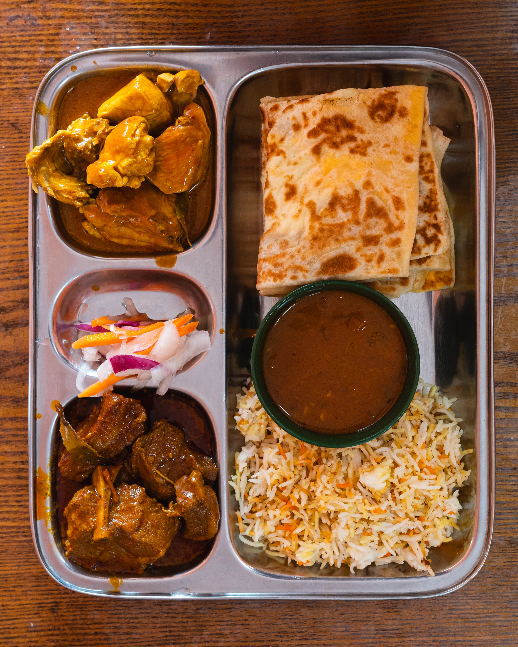 Top down shot of stainless steel tray with curries, pickled vegetables, briyani and prata