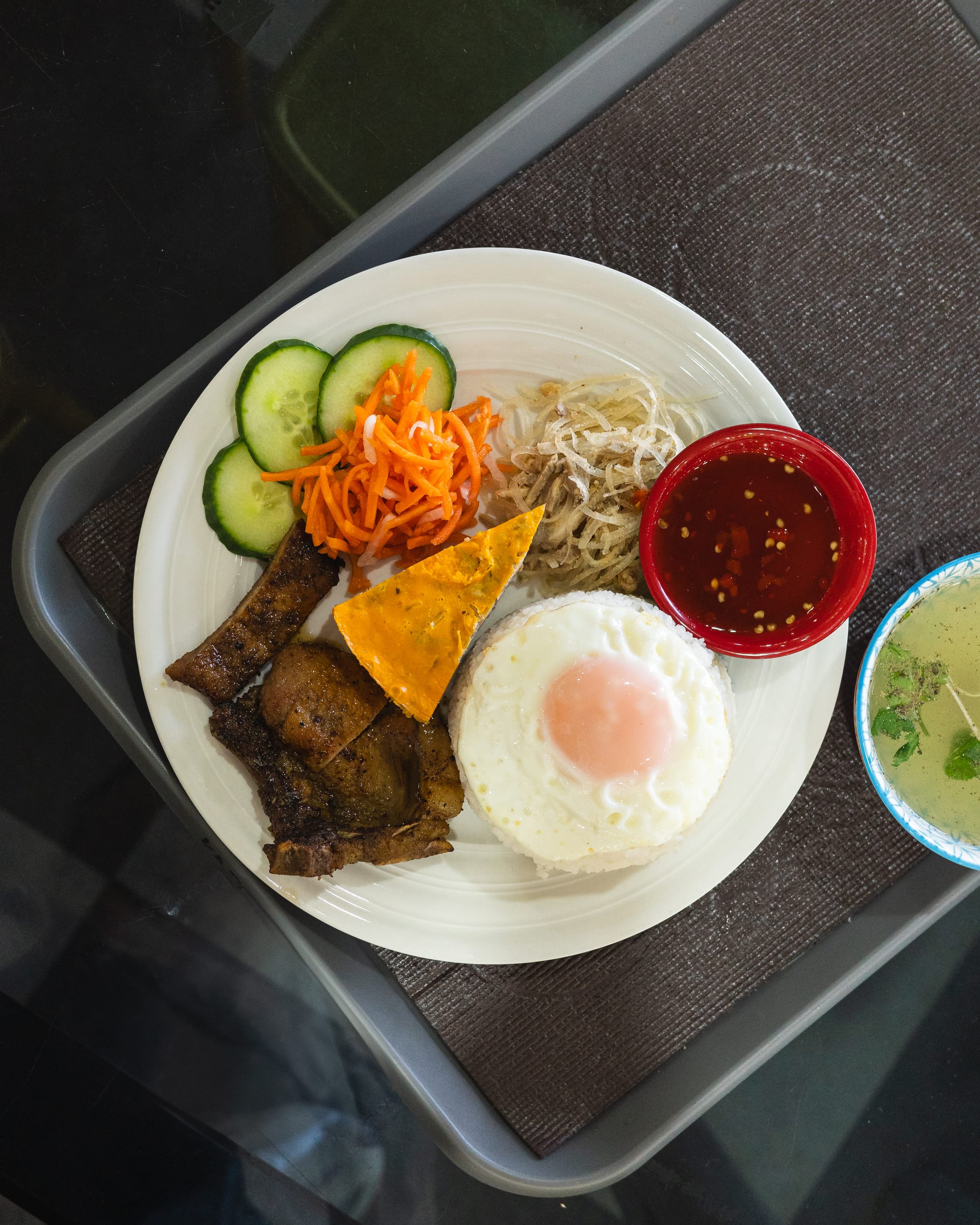 Top down shot of broken rice with pork chop, steamed egg, shredded pork, fried egg, fish sauce, cucumber, pickles and soup