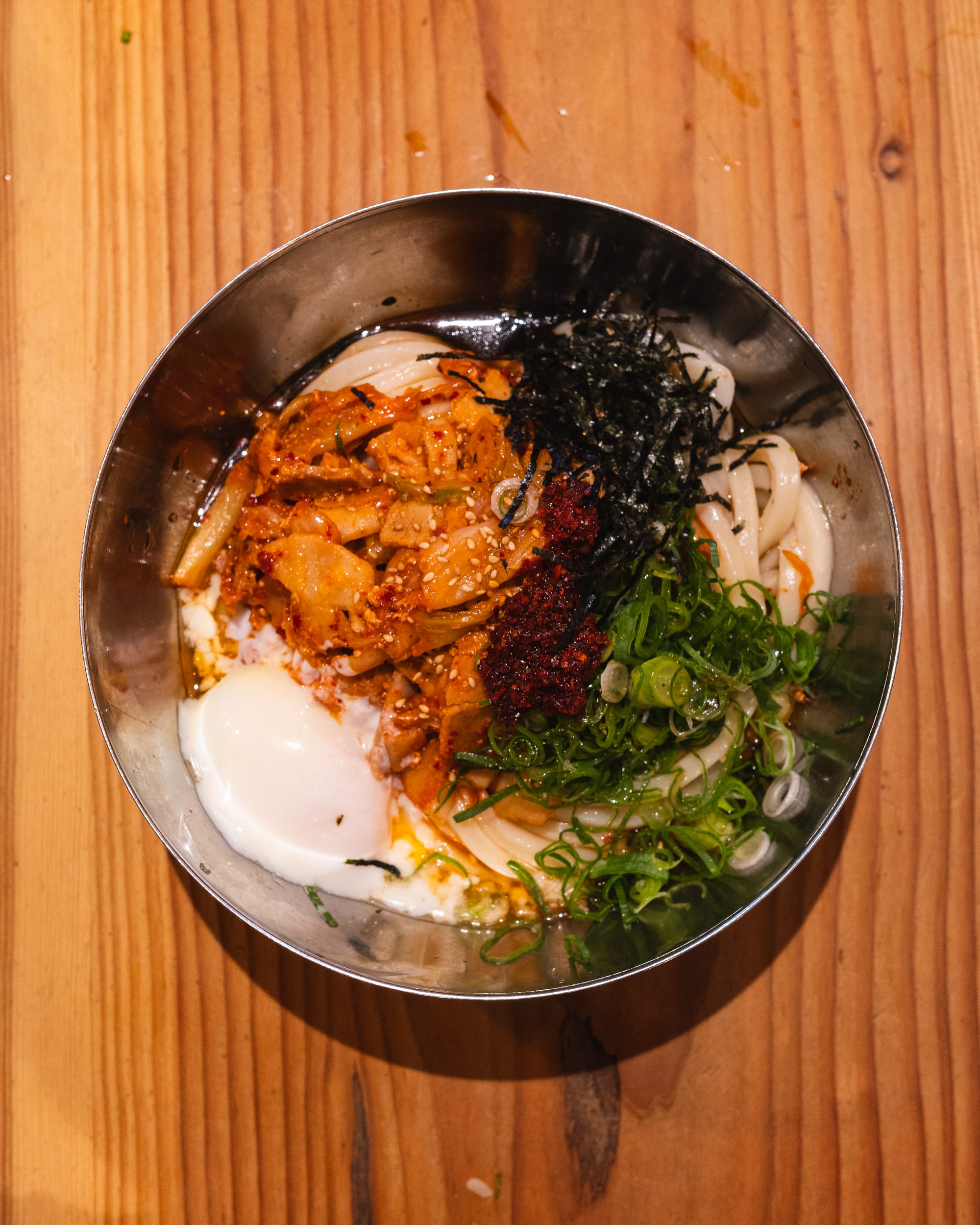 Top down shot of steel bowl with kimchi, onsen egg, chilli crunch, spring onions, seaweed and udon