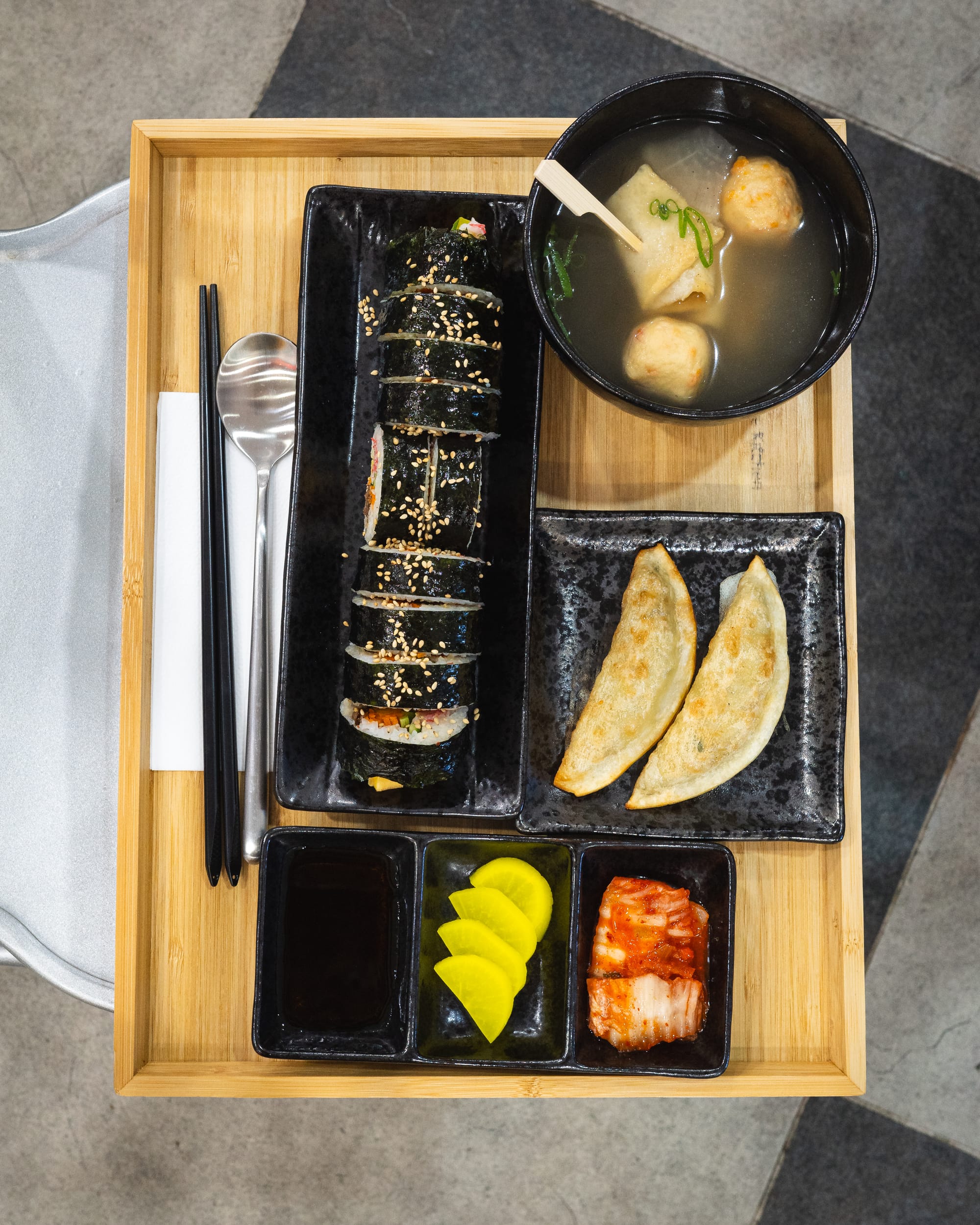 Top down shot of kimbap on a tray with fried dumplings, soup, kimchi and Japanese pickle