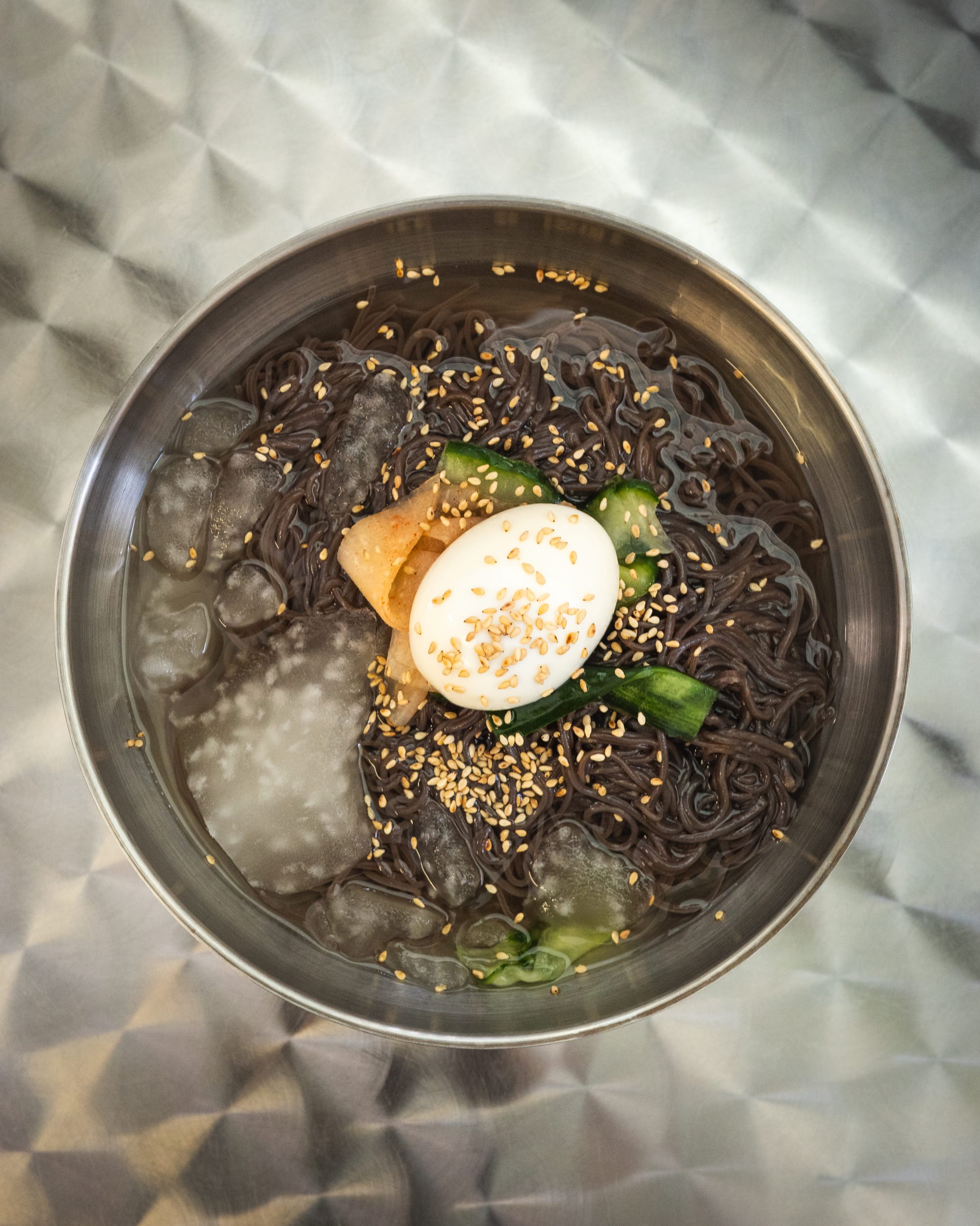 Top down shot of Korean cold noodles with ice, buckwheat noodles, seasame seeds, sliced cucumber and egg