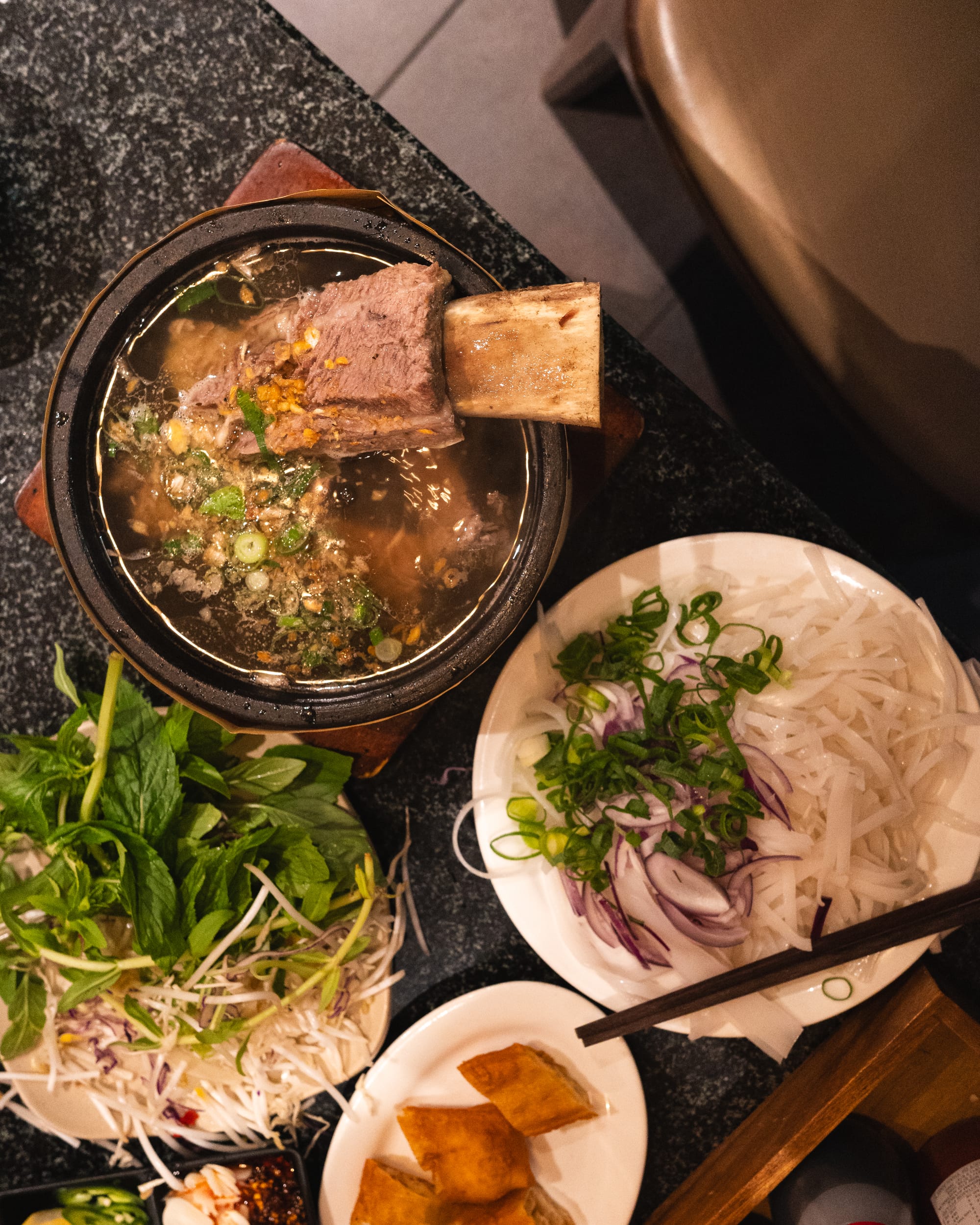 Top down shot of pho in a hot pot with beef rib, salad and raw noodles and garnish on side