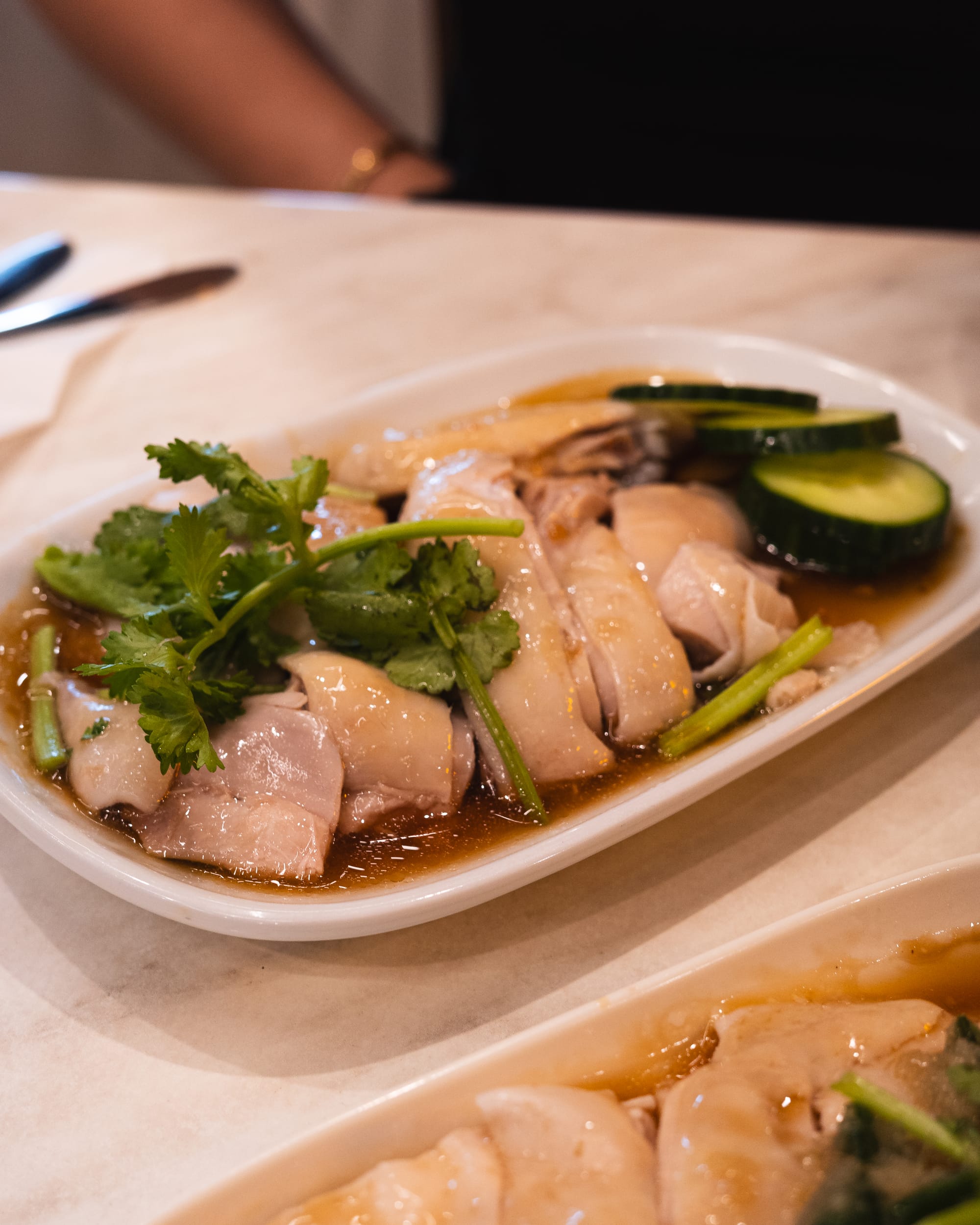 A plate of Hainanese chicken served with soy sauce and garnished with cilantro and cucumber slices.