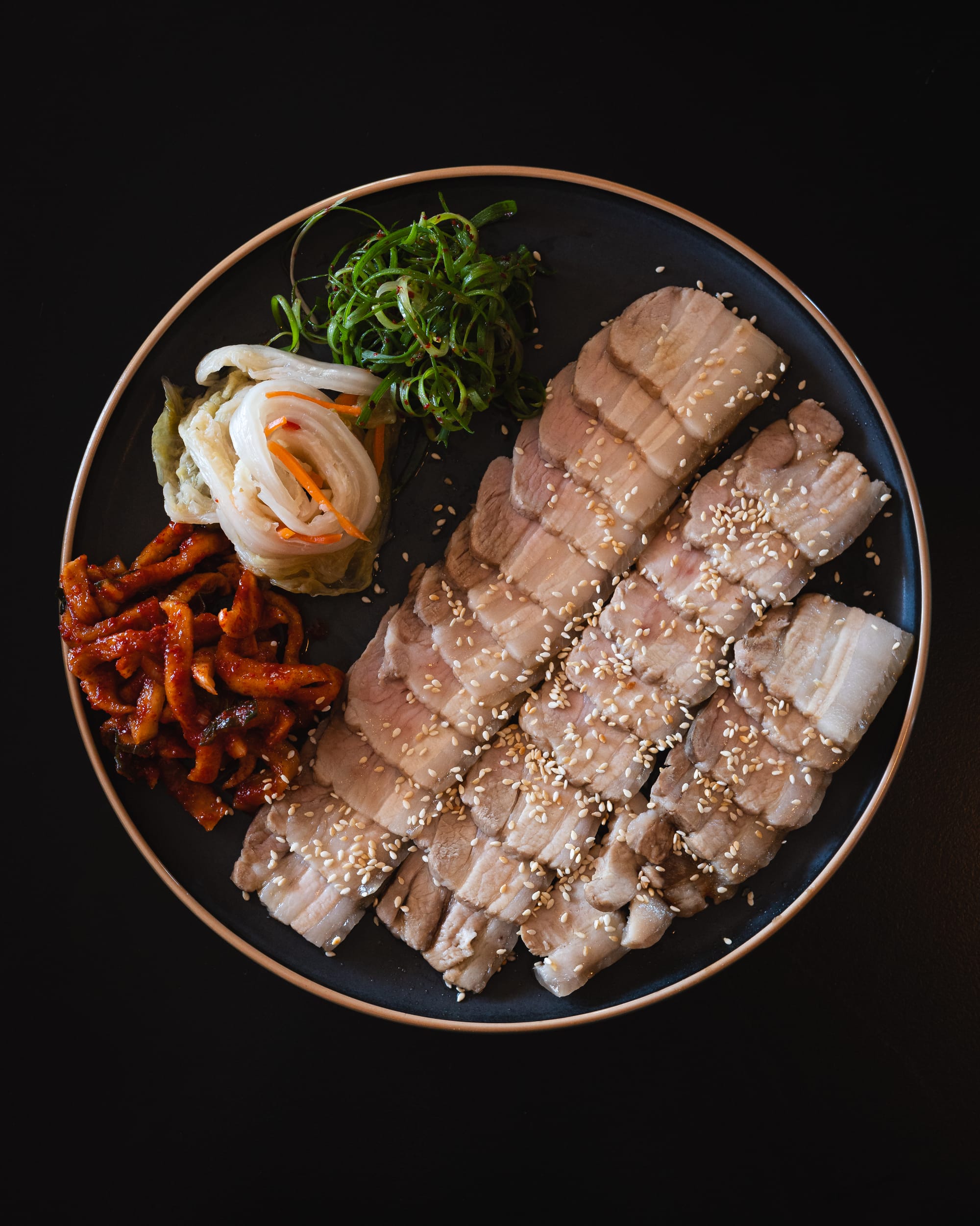 A plate of sliced pork belly sprinkled with sesame seeds and served with pickled vegetables.