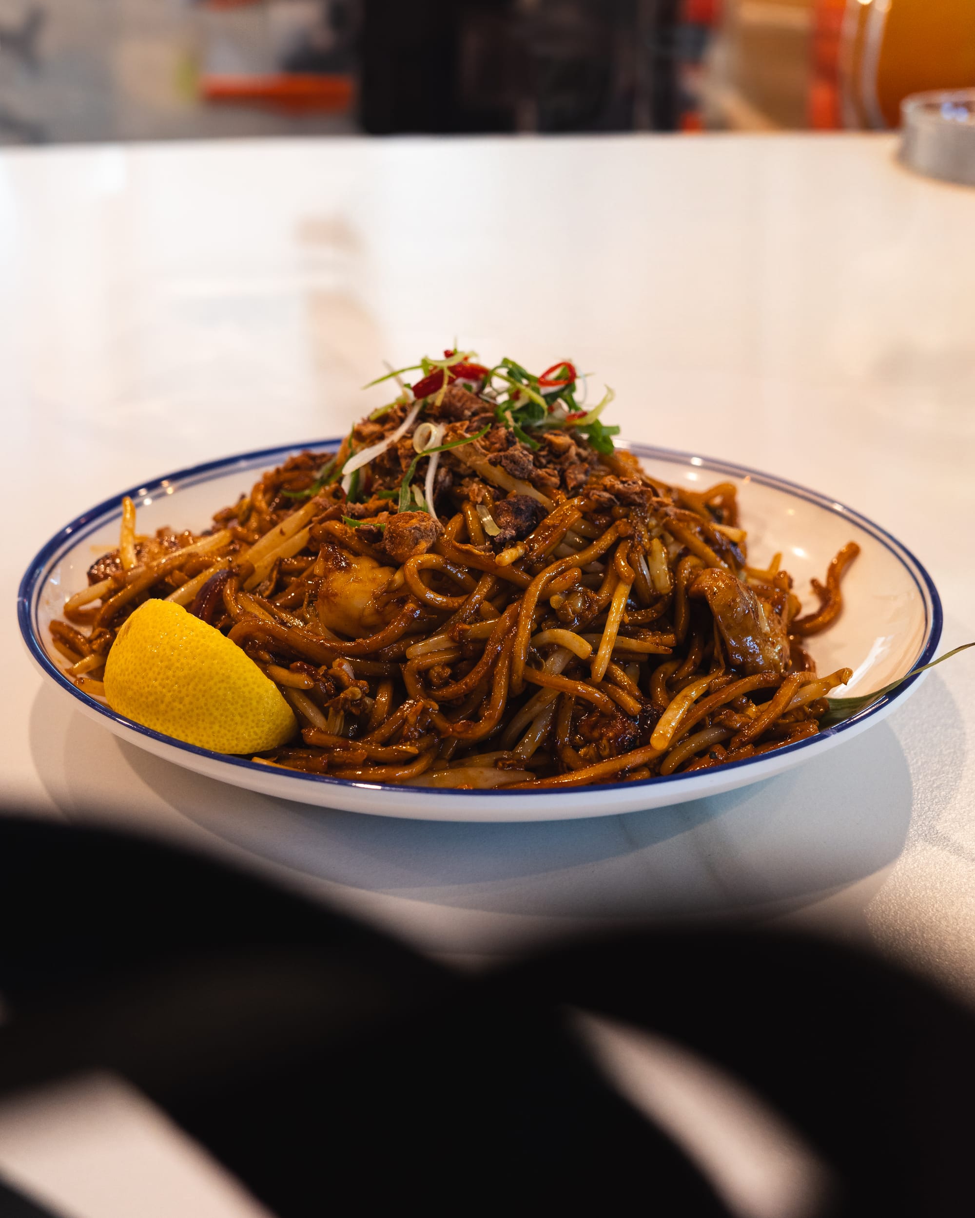 A bowl of stir-fried noodles garnished with fresh green onions and a wedge of lemon.