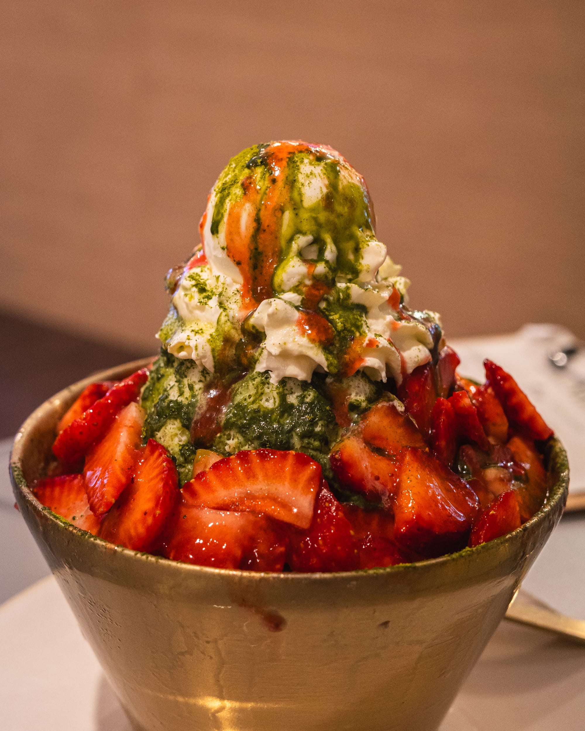 A bowl of strawberry bingsu topped with matcha ice cream, condensed milk, and syrup.