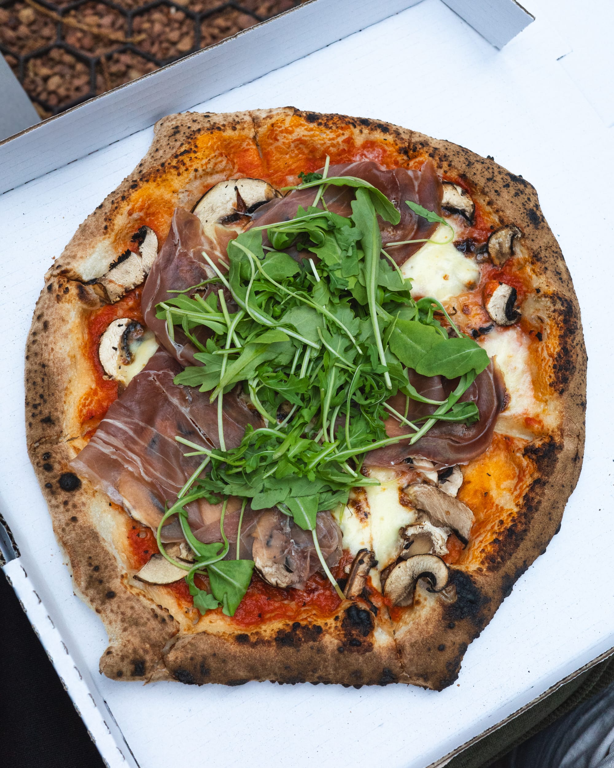 A prosciutto and rocket pizza with a crispy crust in a white box.