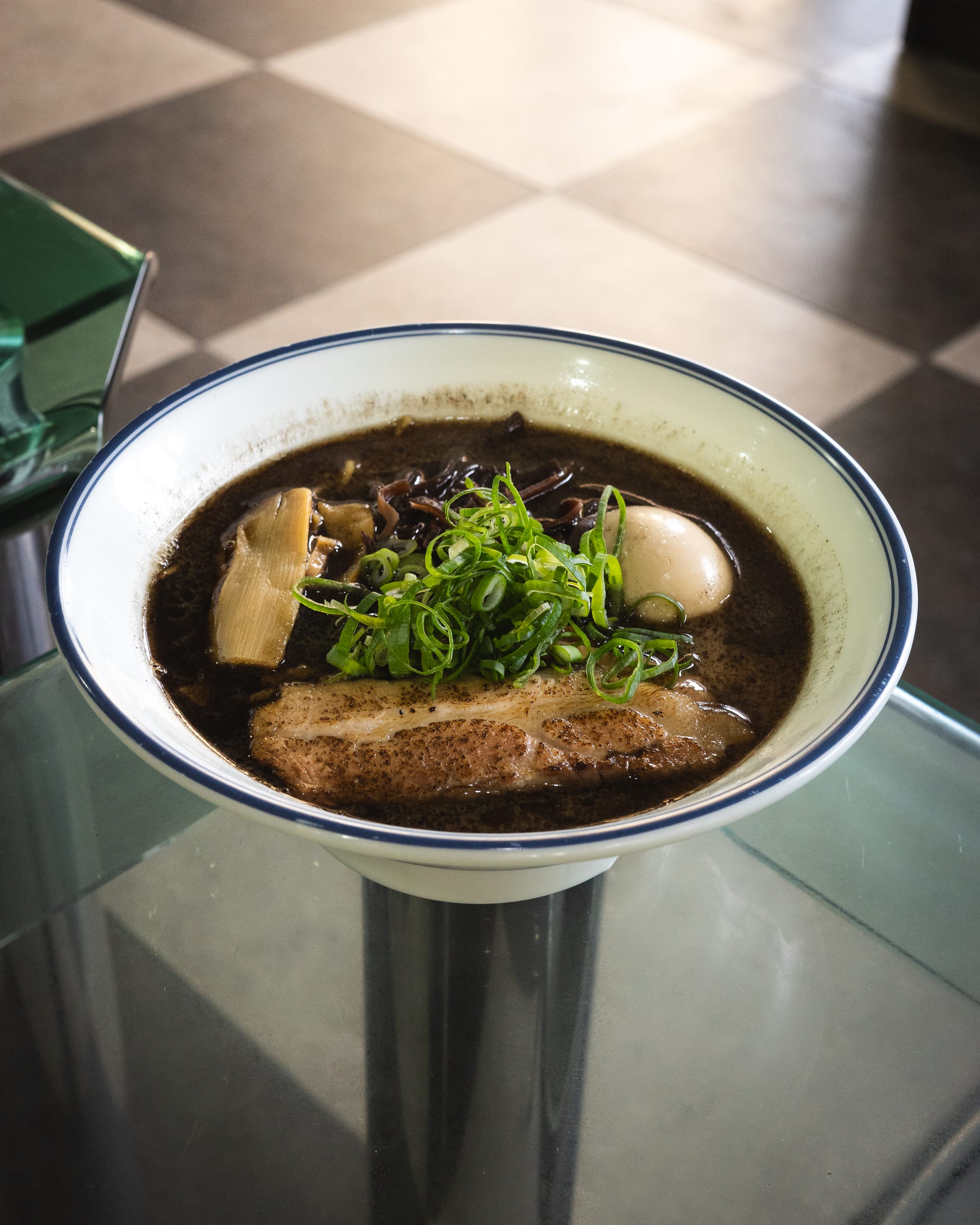 A bowl of ramen with sliced pork, green onions, and mushrooms in a dark-colored broth.