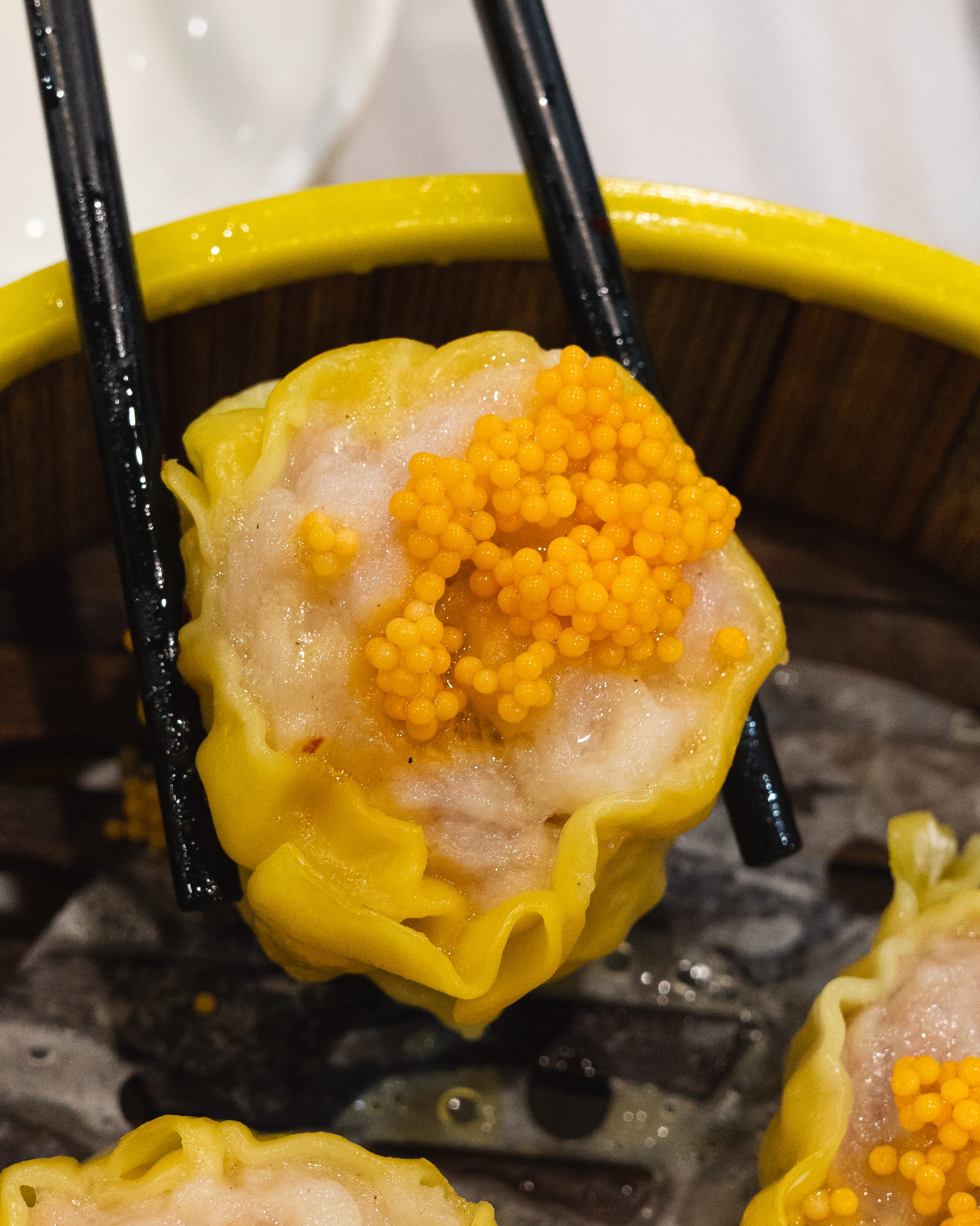 A dim sum dumpling topped with bright orange fish roe, held by black chopsticks.