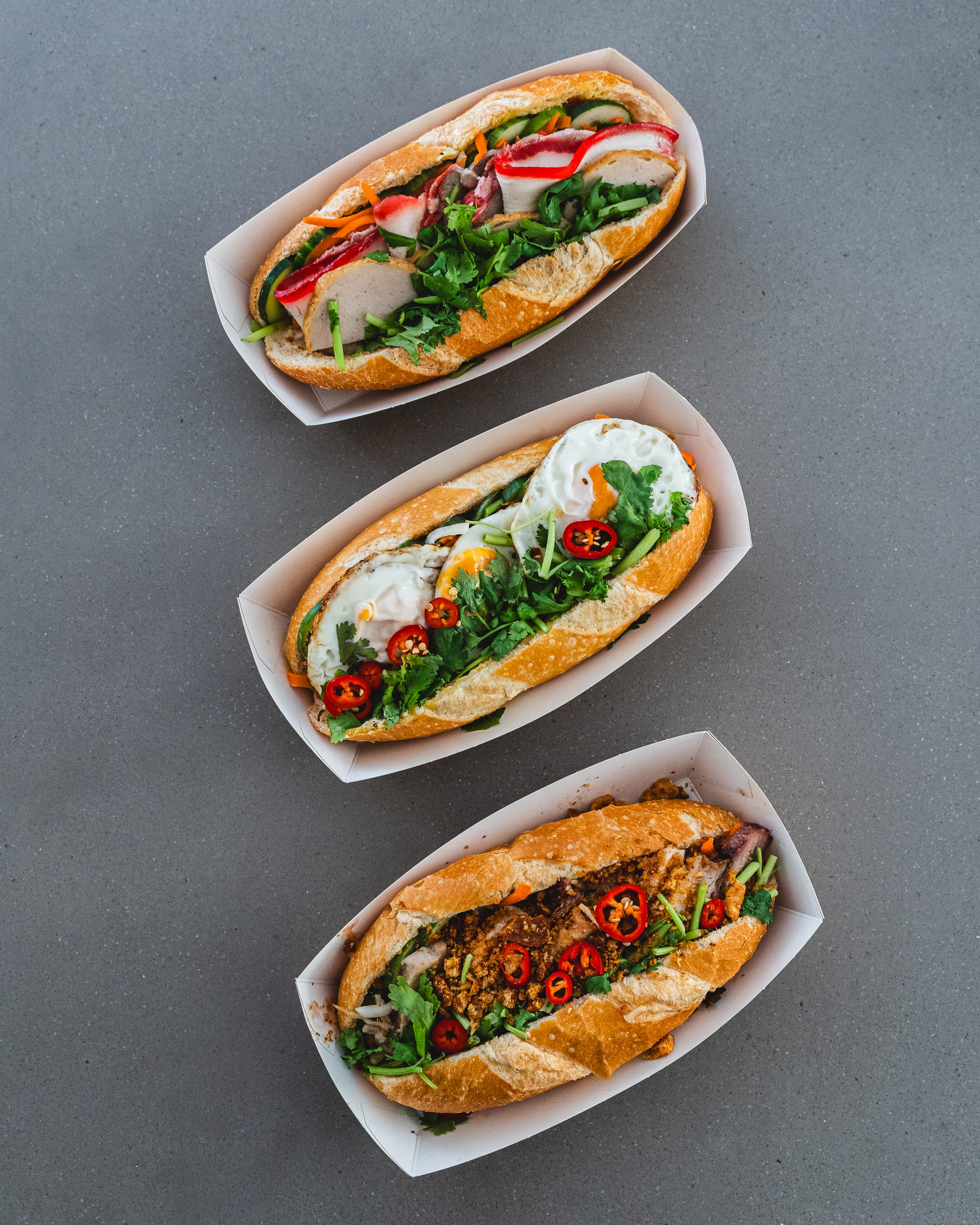 A slate platter with three banh mi sandwiches, filled with ingredients and garnished with herbs.