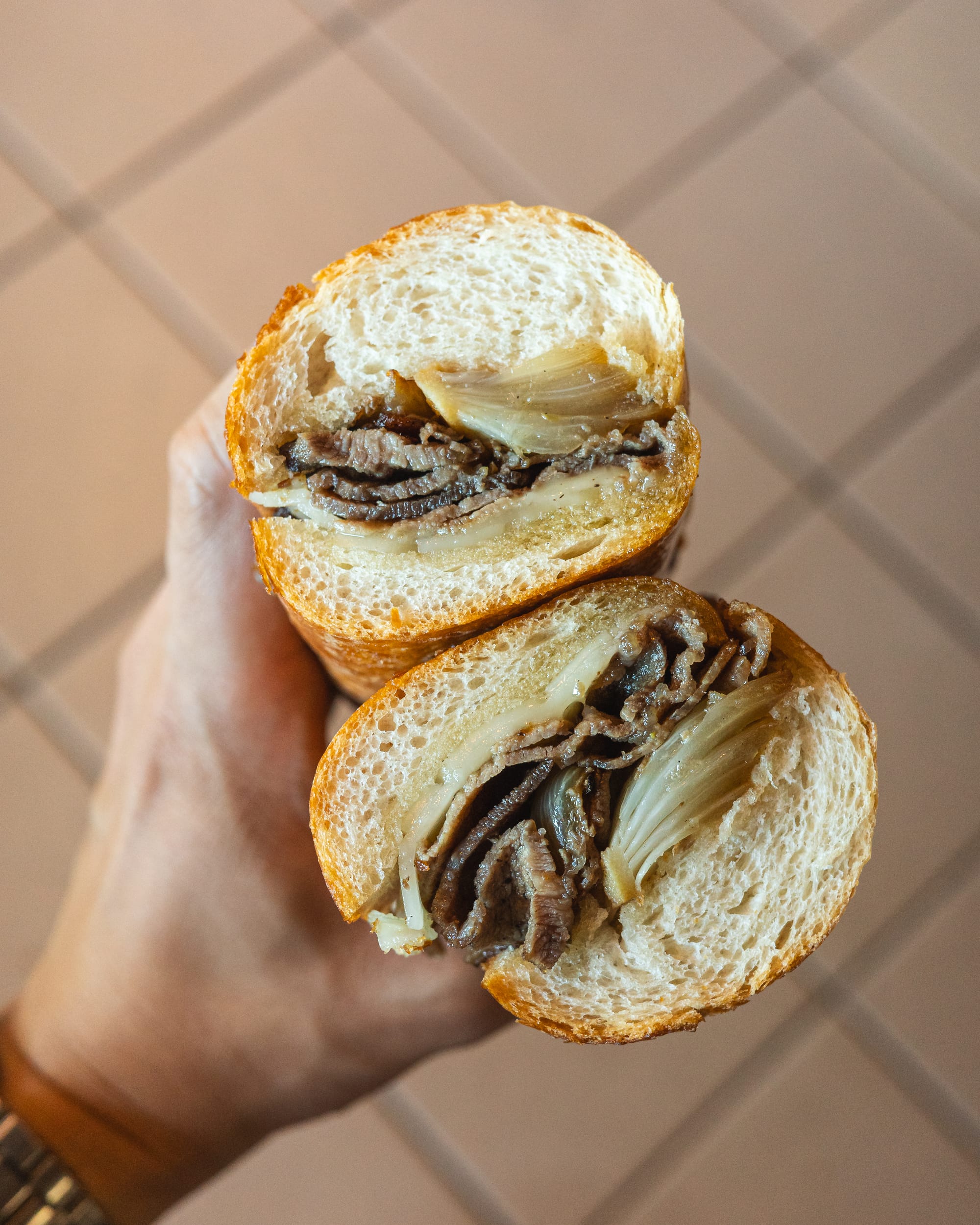 A hand holding a sandwich with sliced roast beef, cheese, and sauce, cut in half.