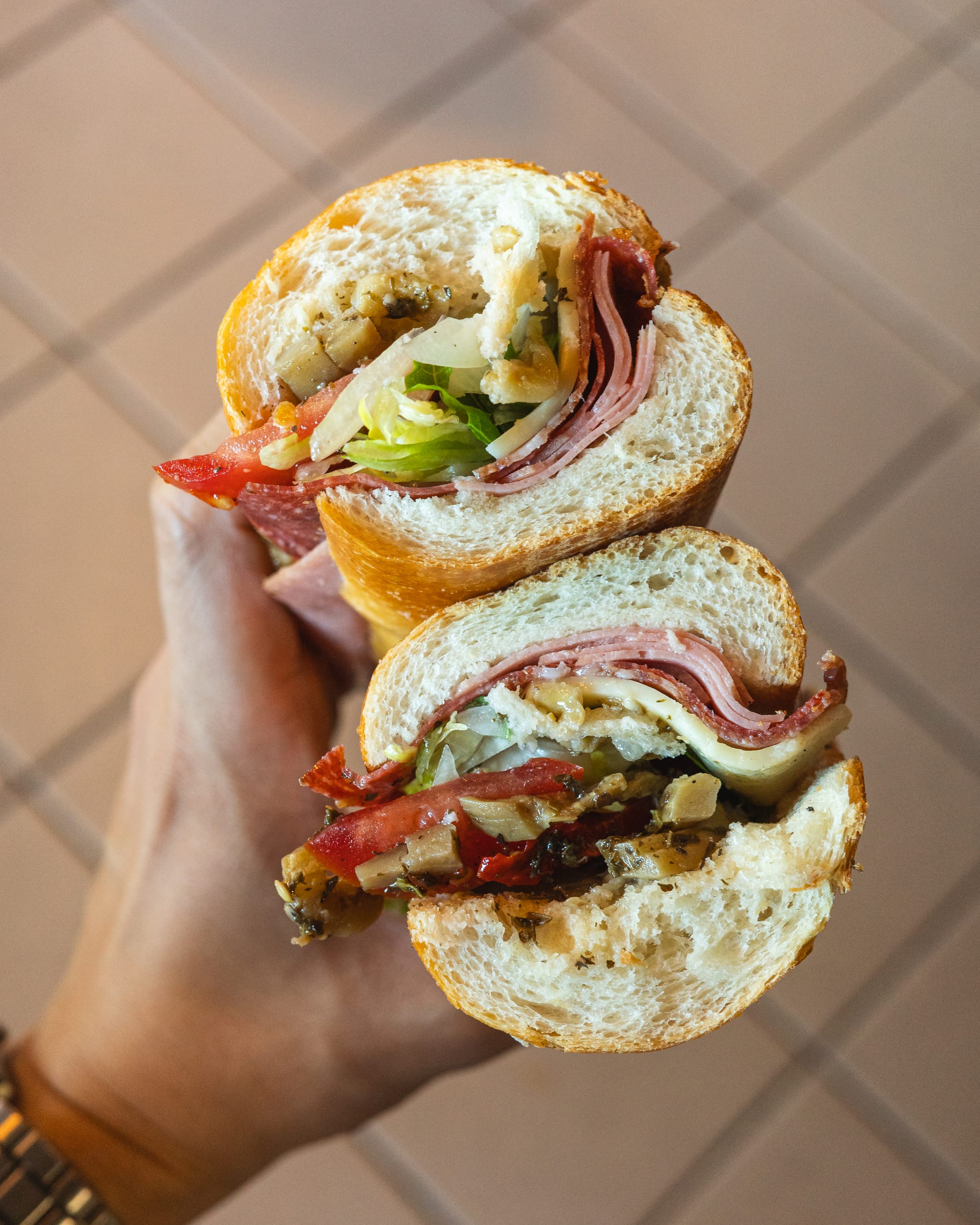 A hand holding a sub-style sandwich with deli meats, cheese, and vegetables.