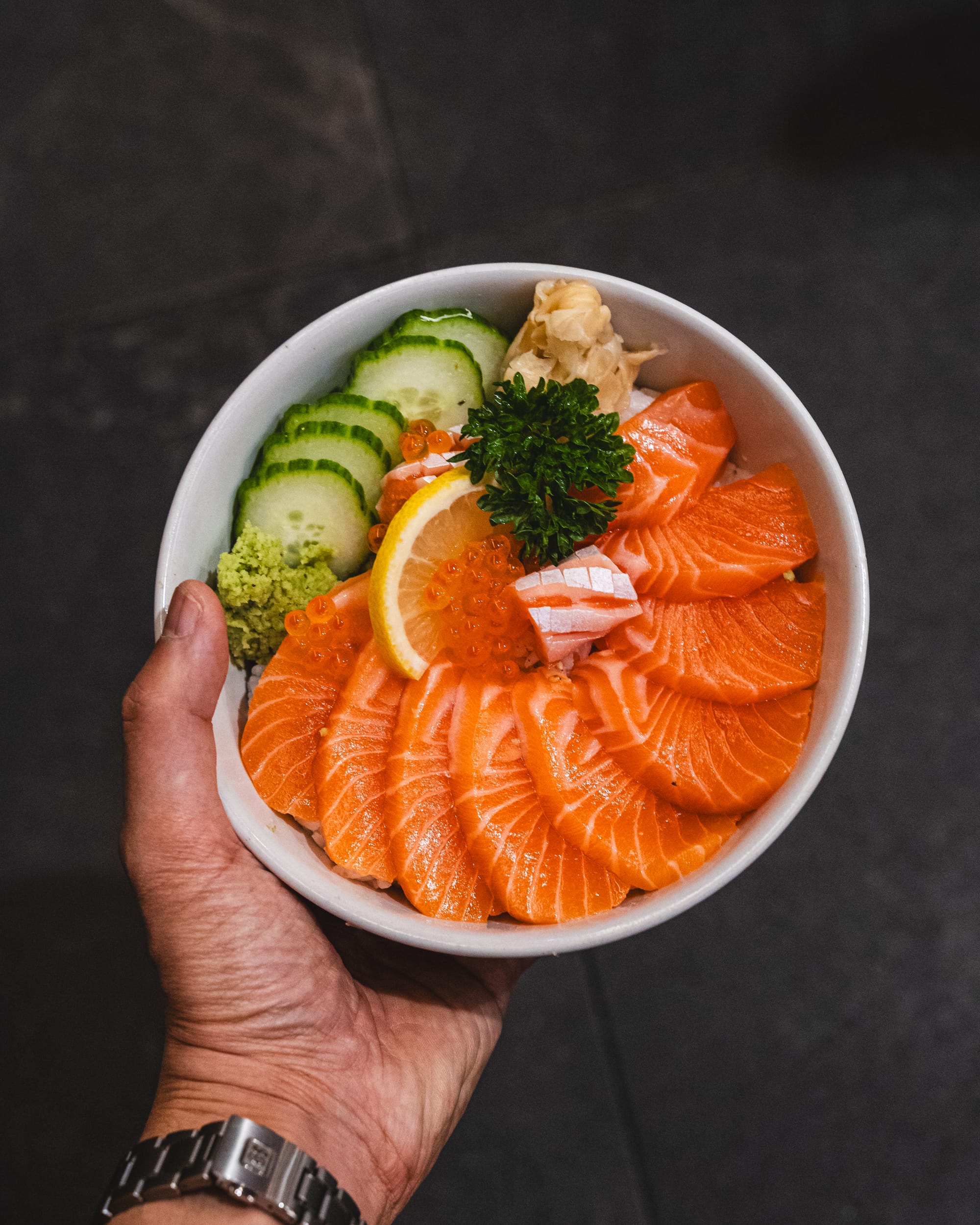 A hand holding a bowl with salmon sashimi, cucumbers, and avocado.